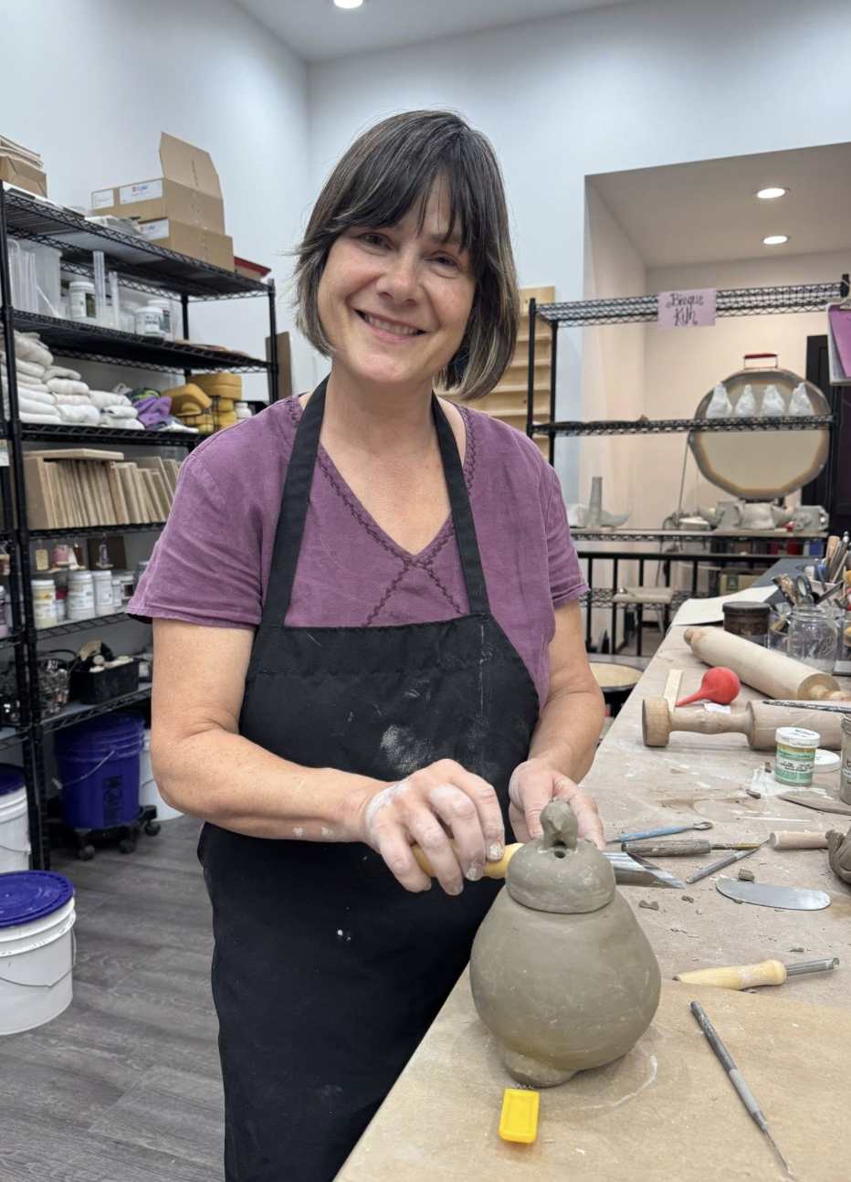 A woman with short brown hair and a purple shirt, smiling while working on a clay pot in a pottery studio.
