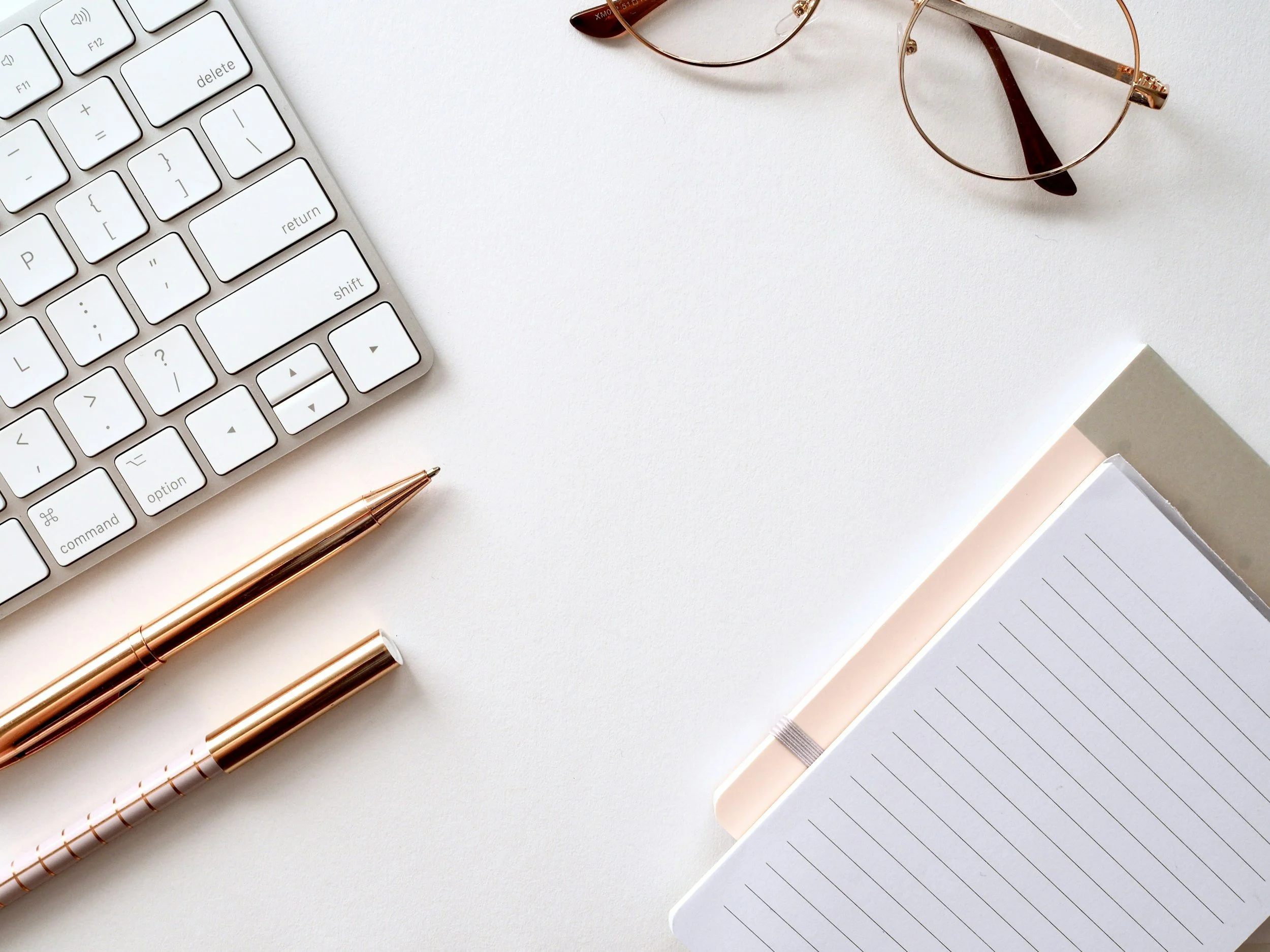 White desk with a partial computer keyboard, pair of sunglasses, two rose gold pens, and an open notebook with lined paper.