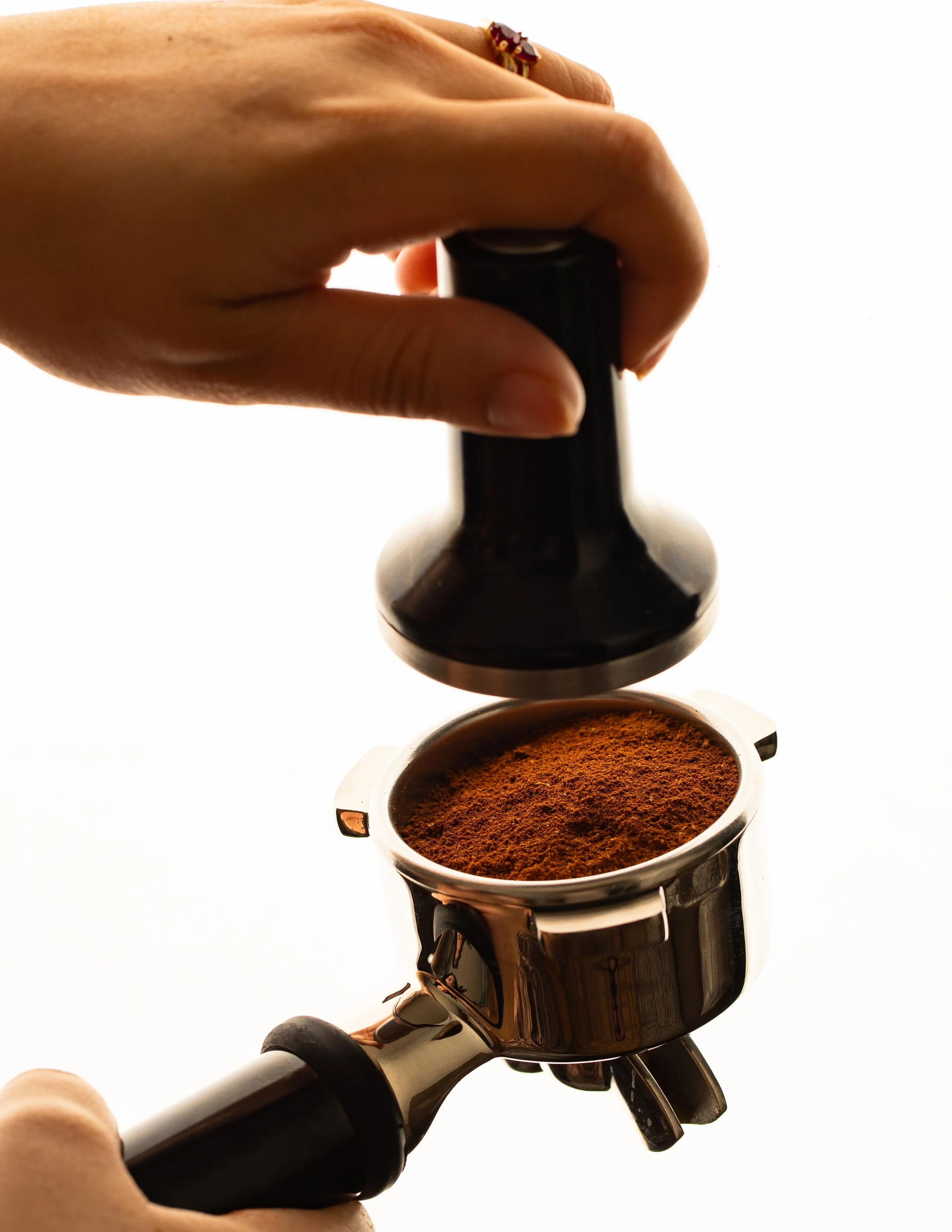 Close-up of a hand holding a portafilter filled with ground coffee, with a tamping tool pressing down on the coffee, set against a white background.