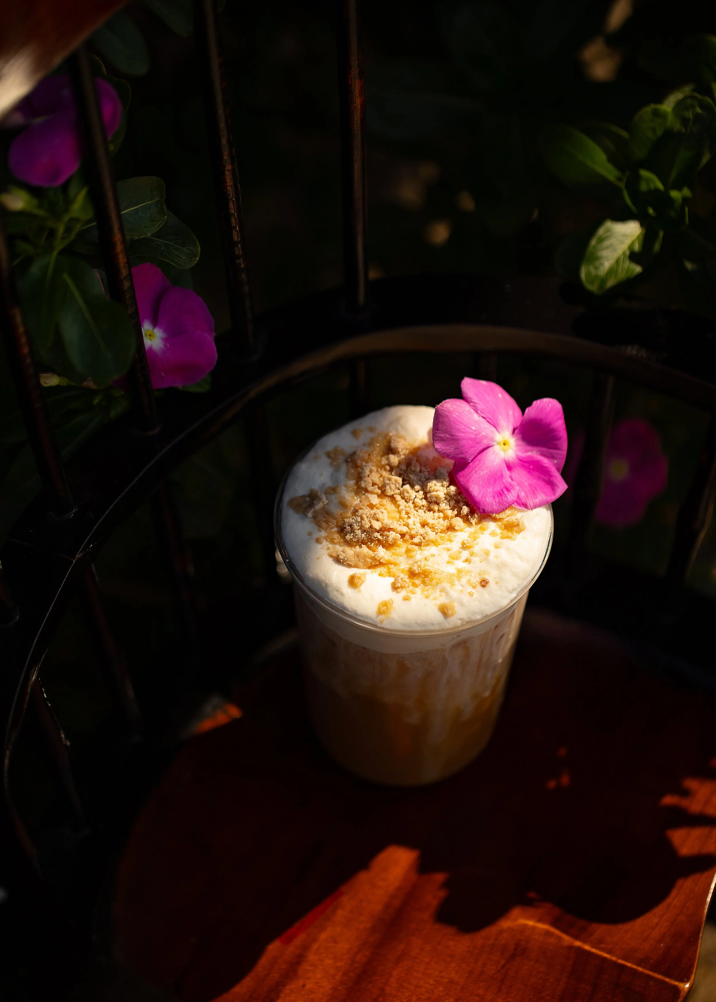 A creamy beverage topped with crushed cookies and a pink flower resting on the rim, in a glass, set on a dark wooden surface with a decorative metal structure and pink flowers in the background.