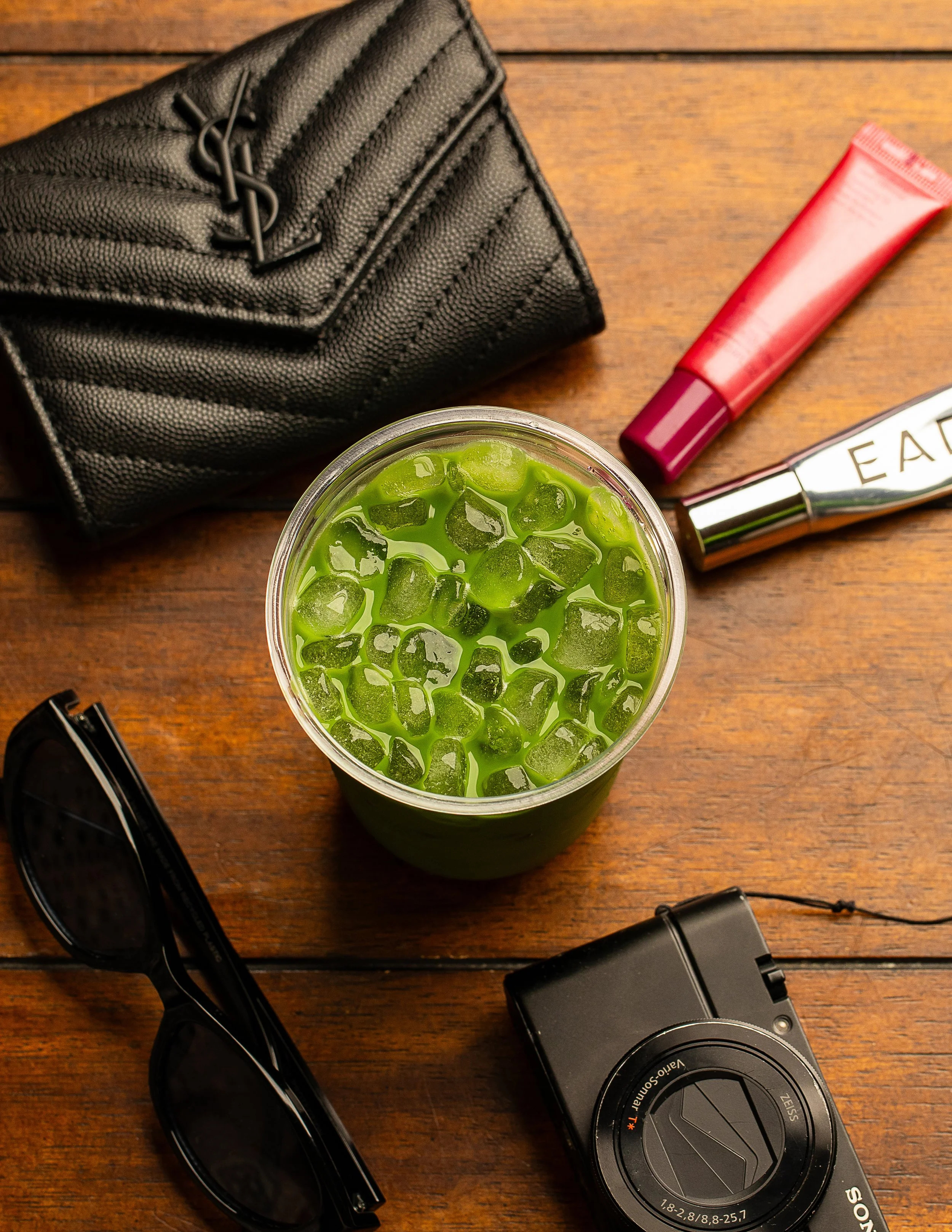 A wooden table with a black purse, a red tube of lotion, a pair of black sunglasses, a camera, and a glass of iced green tea with ice cubes.