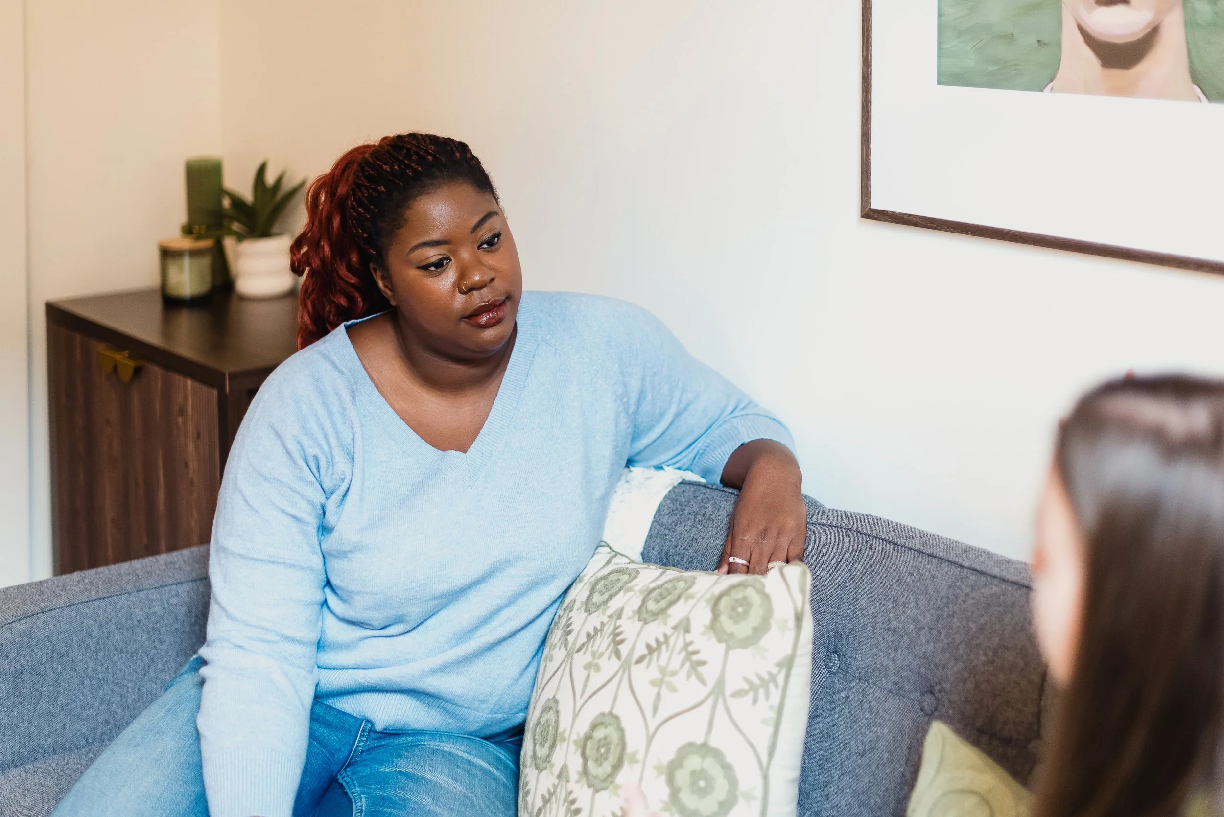 A woman sitting on a gray couch, engaged in conversation with another person, with a wooden side table and a framed picture on the wall behind her.