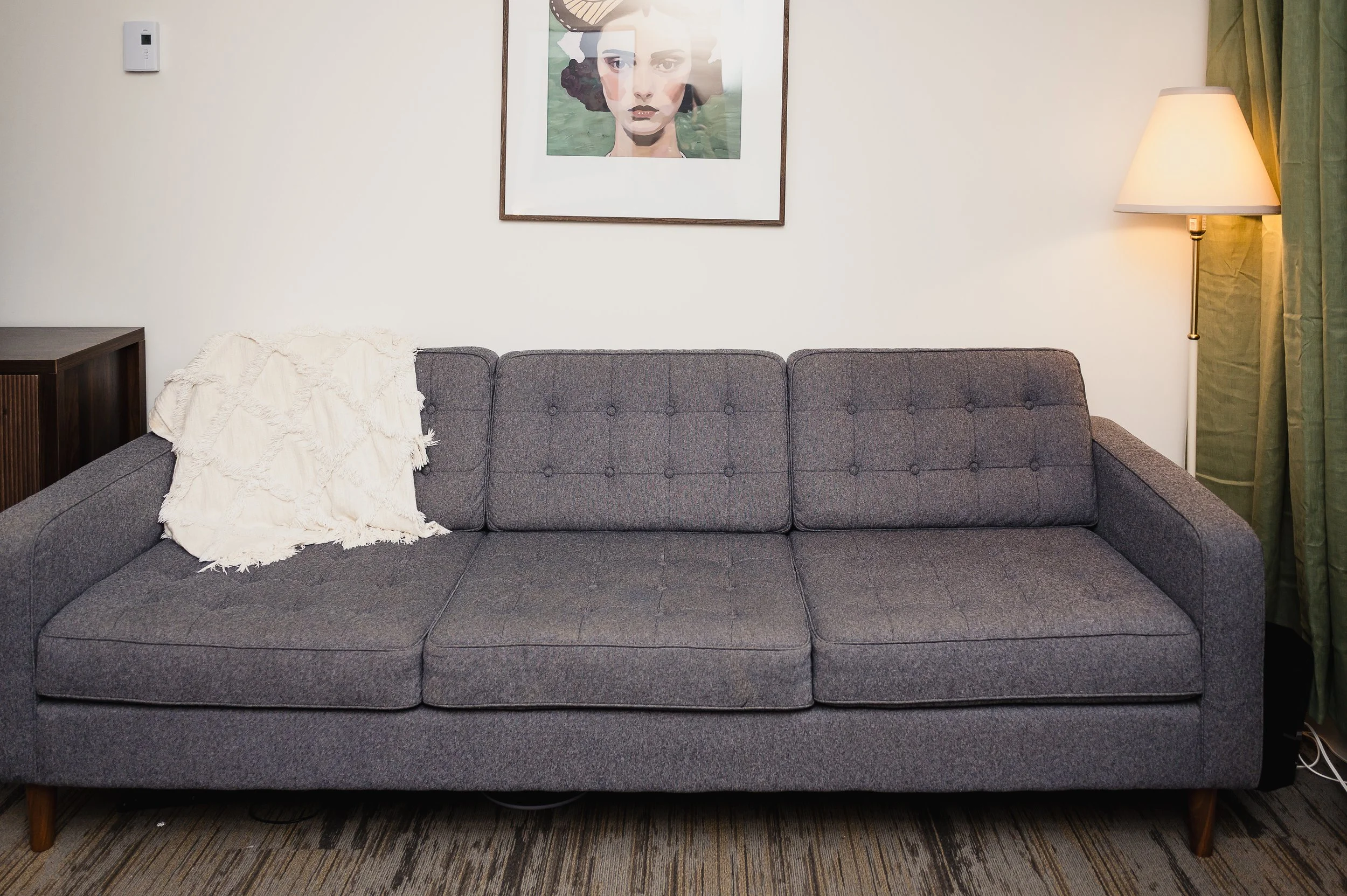 A gray sofa with three seat cushions and a white textured throw blanket on the left armrest, a framed portrait of a woman hanging above, a wooden side table on the far left, a floor lamp with a white shade on the right, green curtains, and a patterned carpet.