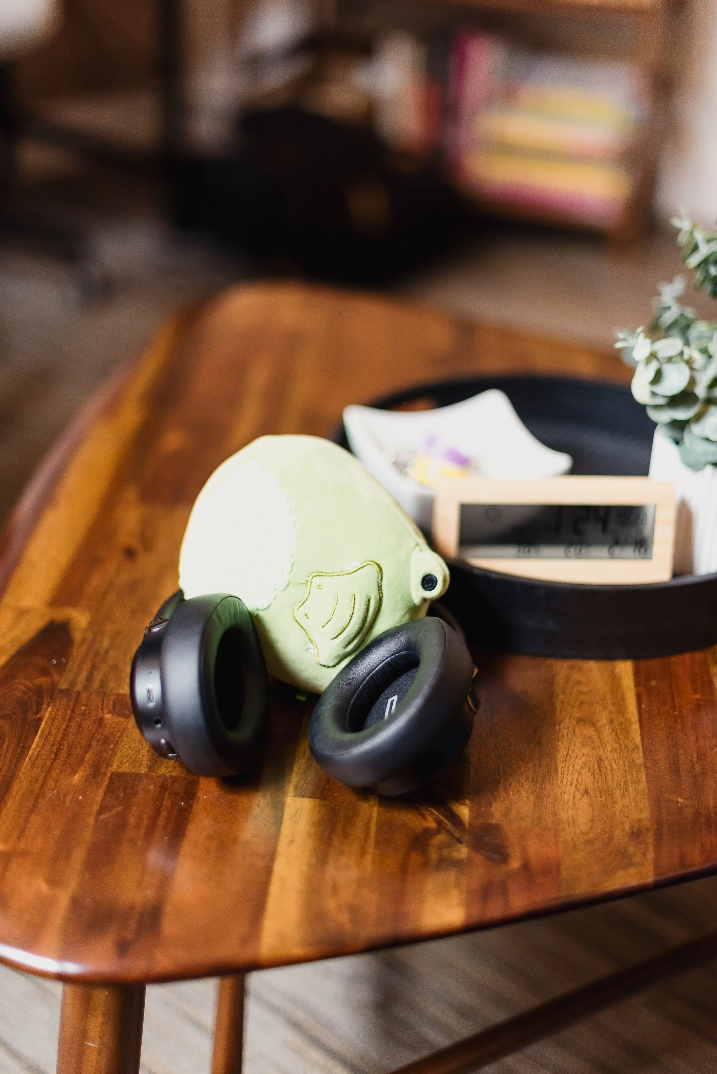 A plush toy shaped like a green chameleon with wing details, a pair of black over-ear headphones, a digital clock displaying "24:14" on a wooden table, with a black tray holding a small dish and a plant, and a bookshelf with colorful books in the background.