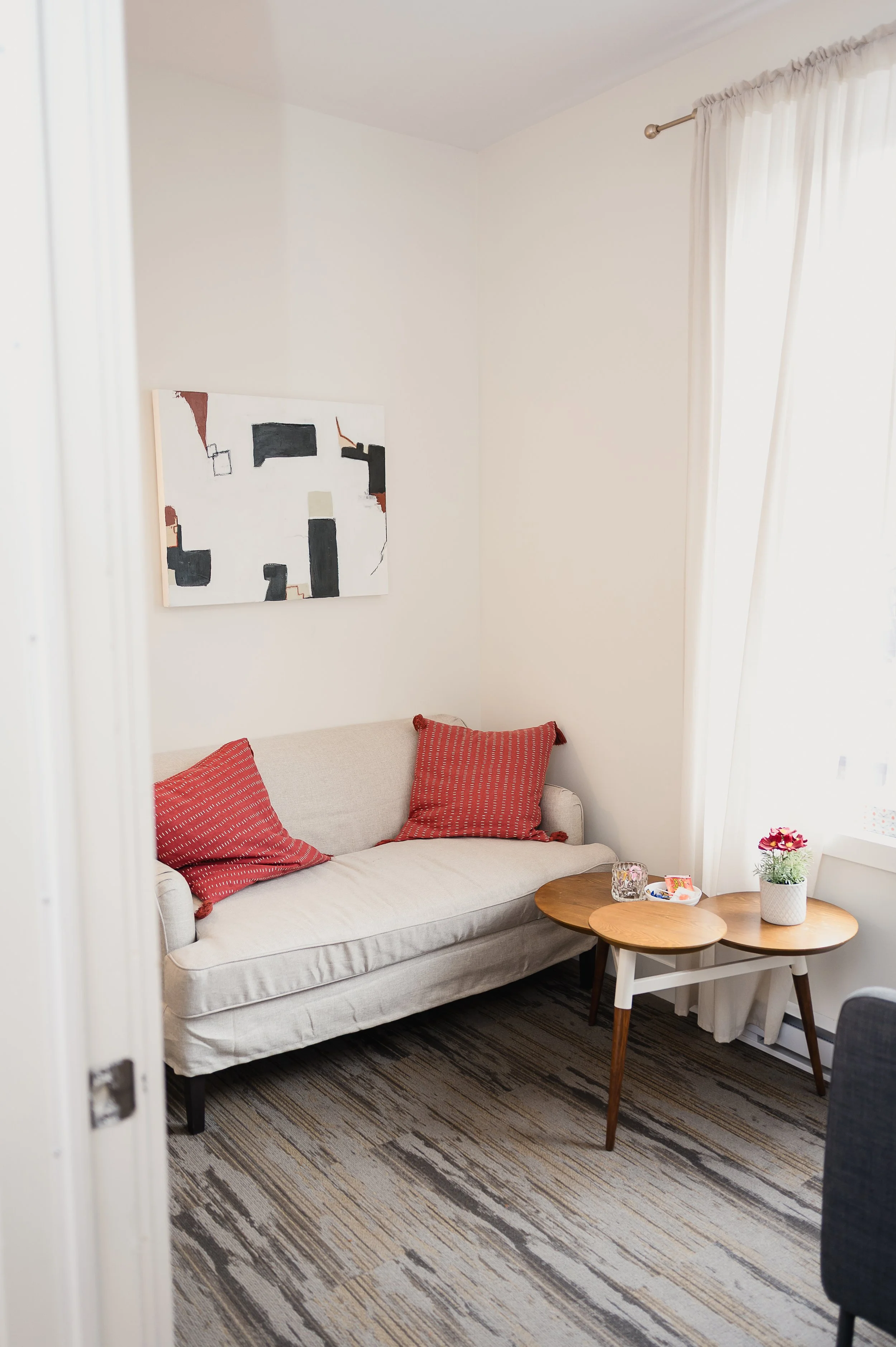 A cozy living room with a white sofa, two red throw pillows, a abstract art piece on the wall, a round wooden side table with a potted plant and glass of water, and a window with white curtains letting in natural light.