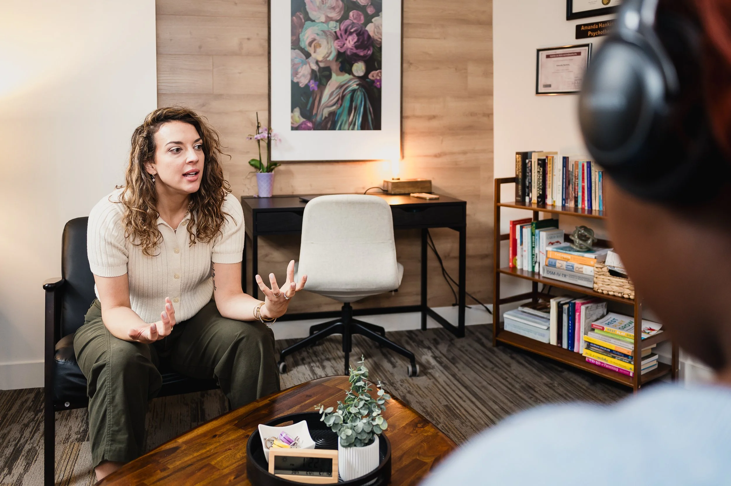 Une femme aux cheveux bouclés assise sur une chaise noire, discutant avec une autre personne portant des écouteurs, dans un bureau douillet ou une salle de thérapie avec un mur en bois, une bibliothèque remplie de livres, un petit bureau avec une orchidée, et une peinture encadrée de fleurs accrochée au mur.
