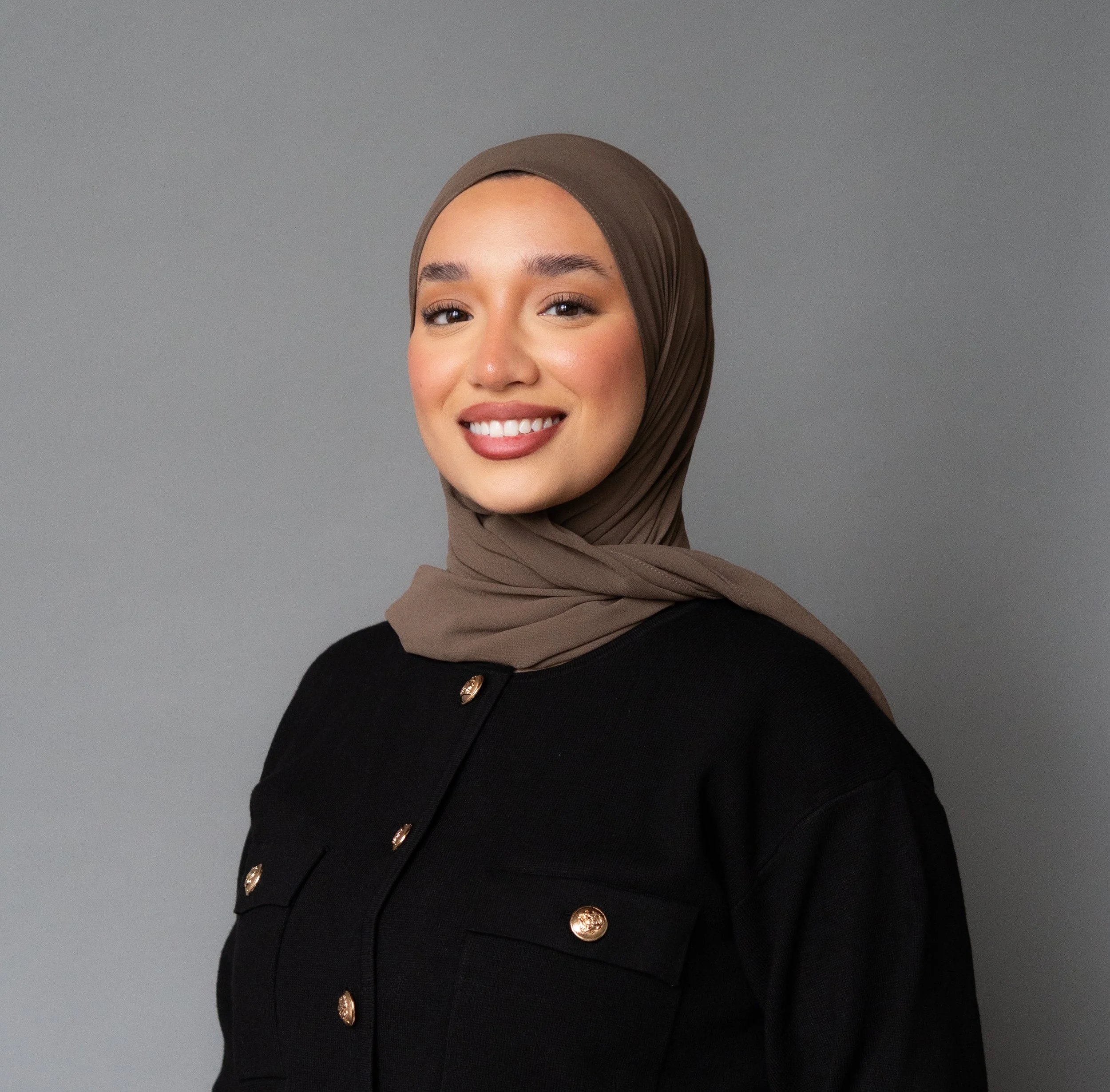 Close-up of a smiling woman wearing a brown hijab and a black top with gold buttons, against a plain grey background.
