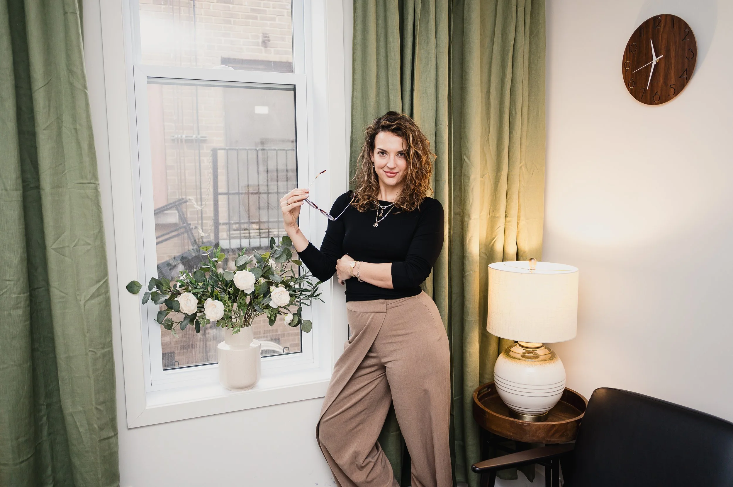 A woman standing next to a window holding sunglasses, with a vase of white flowers, green curtains, a wall clock, a table lamp, and a black chair in the background.