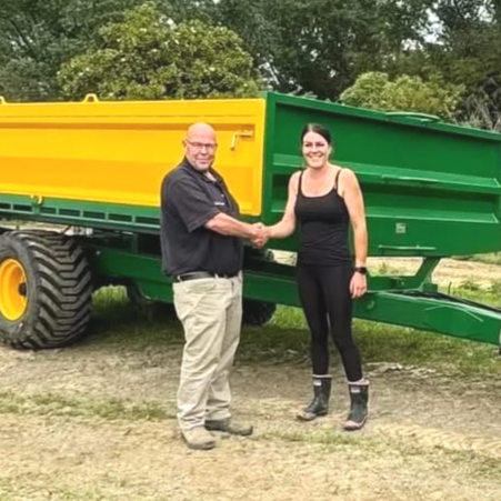 Two people shaking hands in front of a green and yellow tractor and trailer on a farm.
