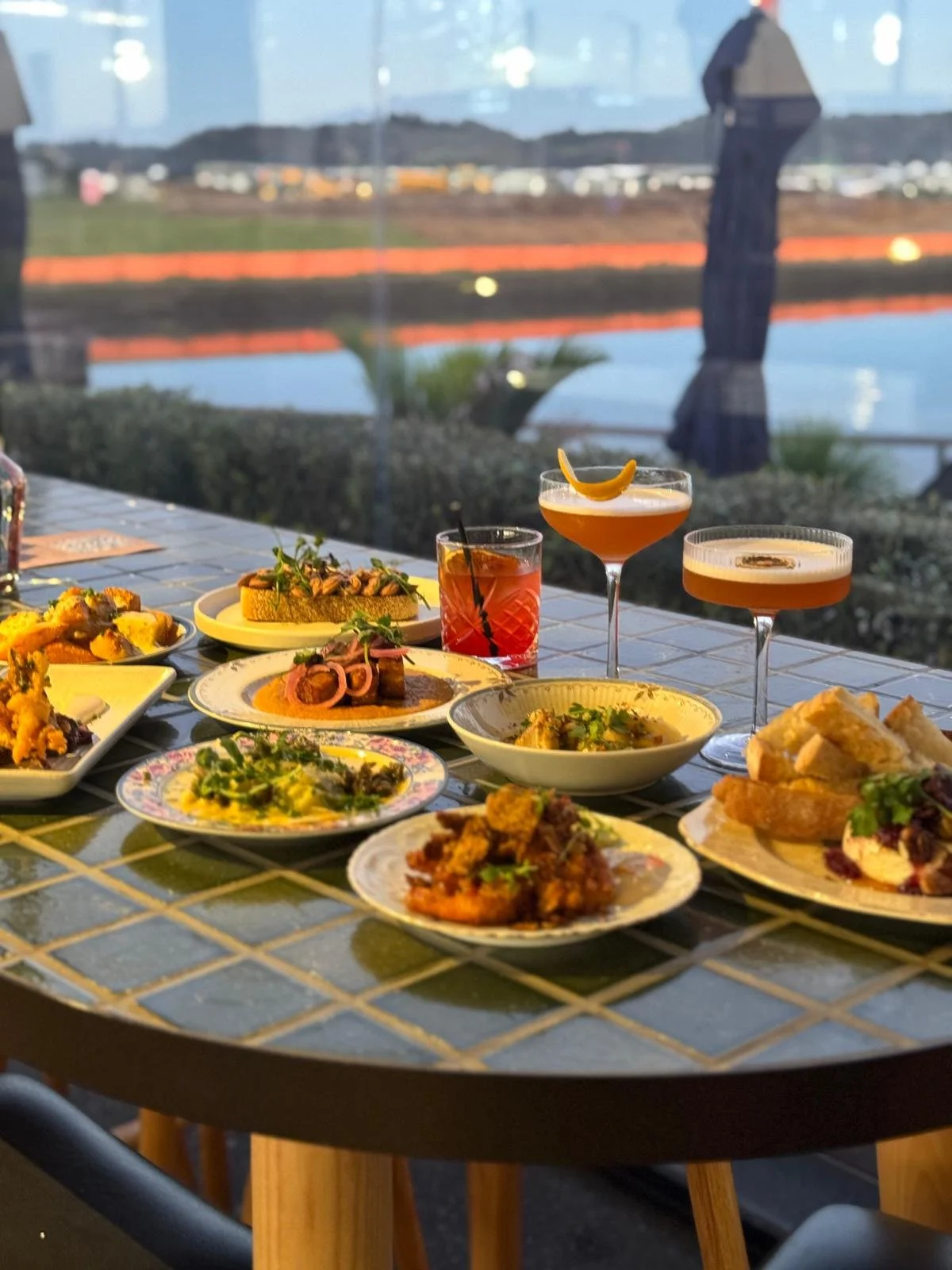 A table set with various dishes and cocktails, located by a large window overlooking a body of water and outdoor umbrellas, with a background of a landscape featuring distant buildings and trees.