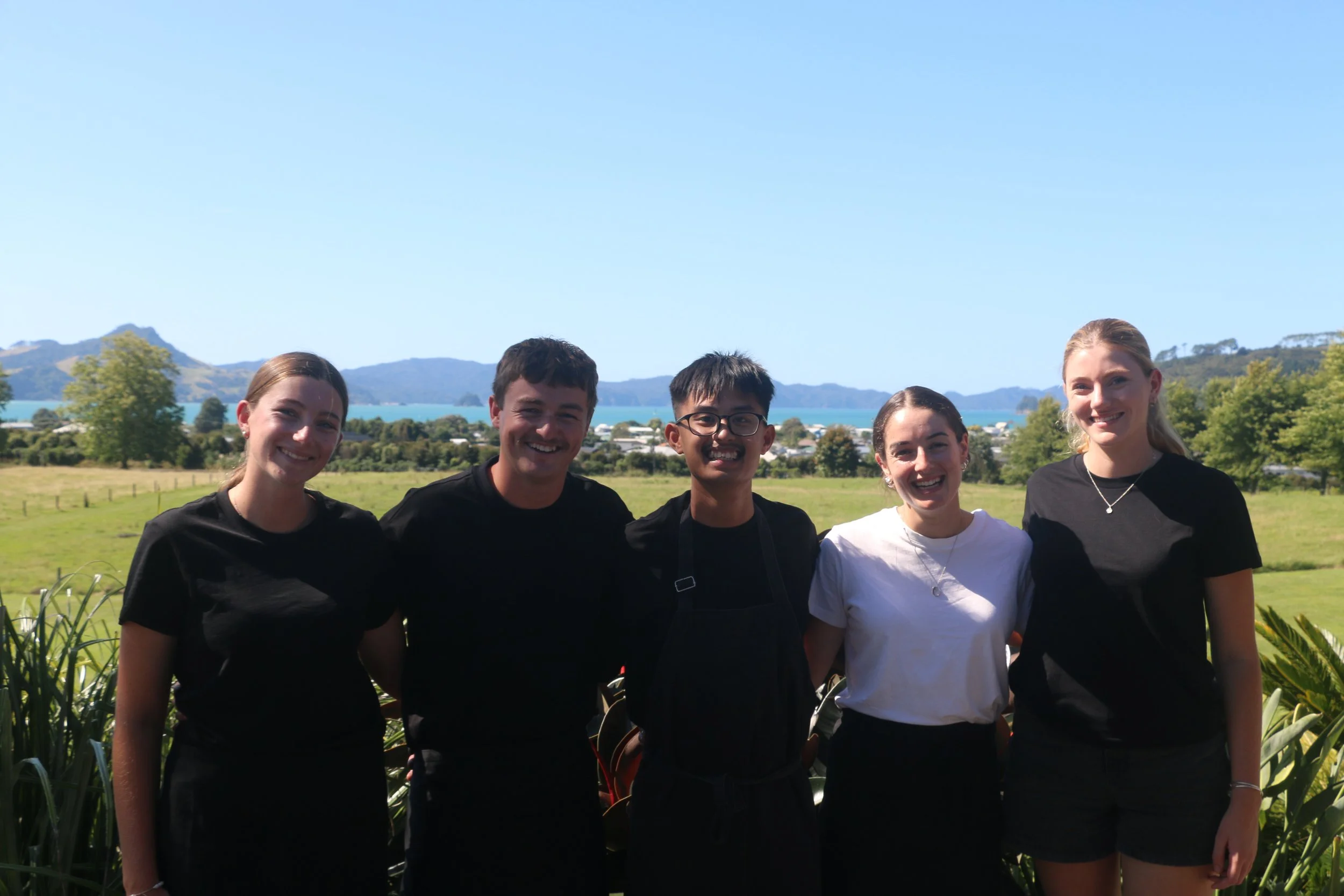 A group of five young people smiling outdoors with a scenic background of green fields, trees, water, and mountains under a clear blue sky.