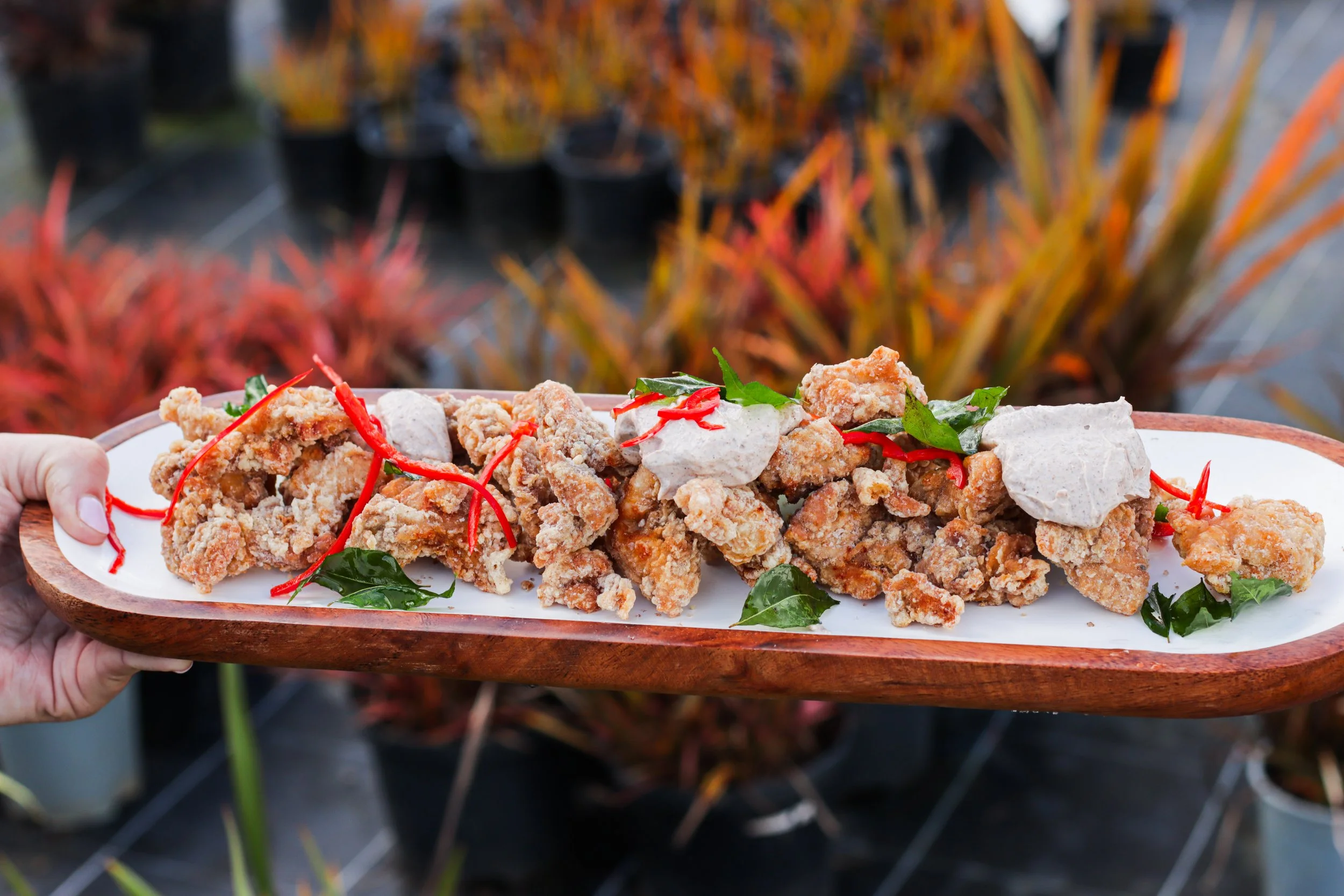 A person holding a wooden tray of fried chicken pieces garnished with red chili slices and green herbs, outdoors with blurred autumn foliage in the background.