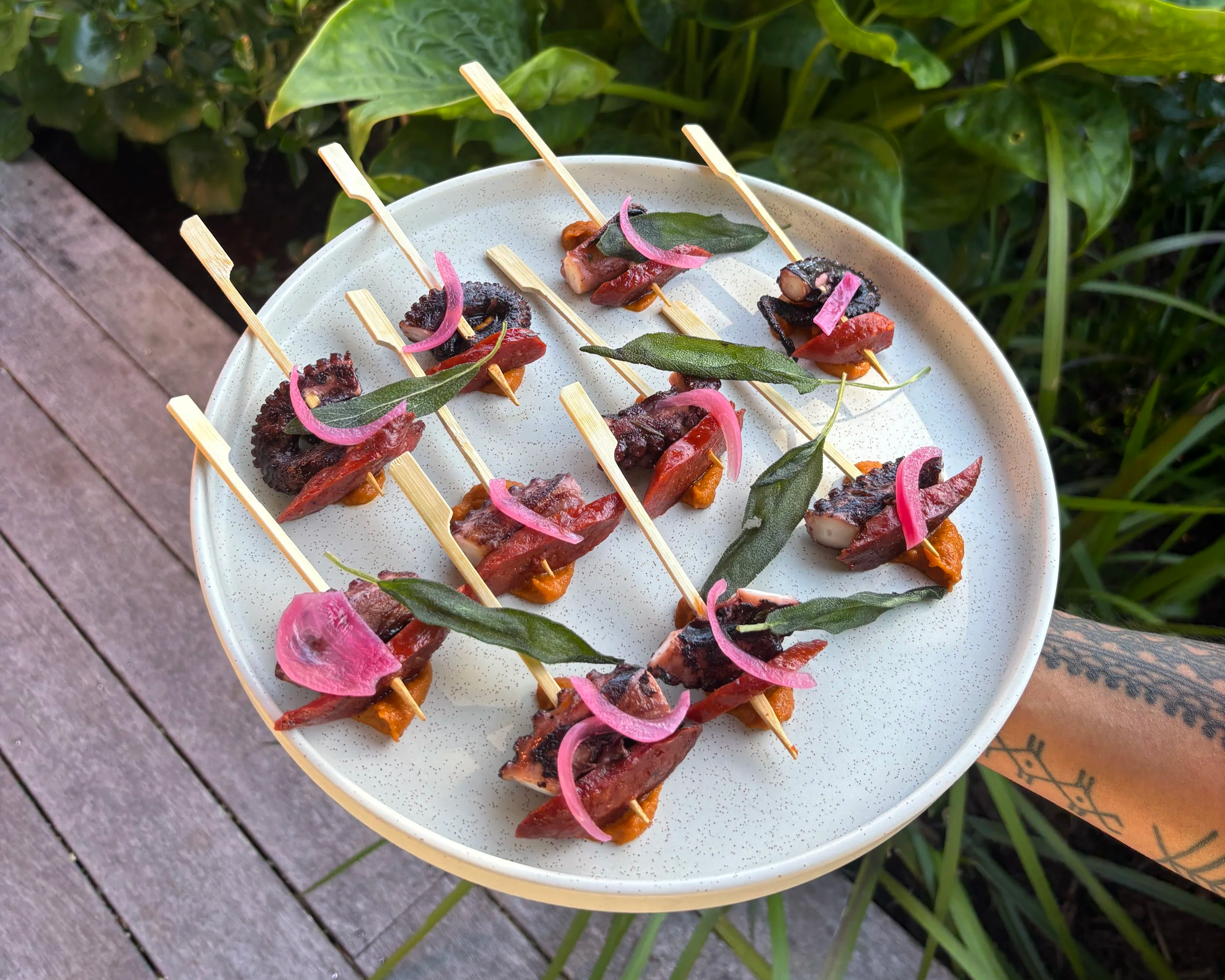 Plate of skewered small appetizers with meat, vegetables, and herbs, garnished with pickled pink onions, set outdoors on a table with green foliage in the background.