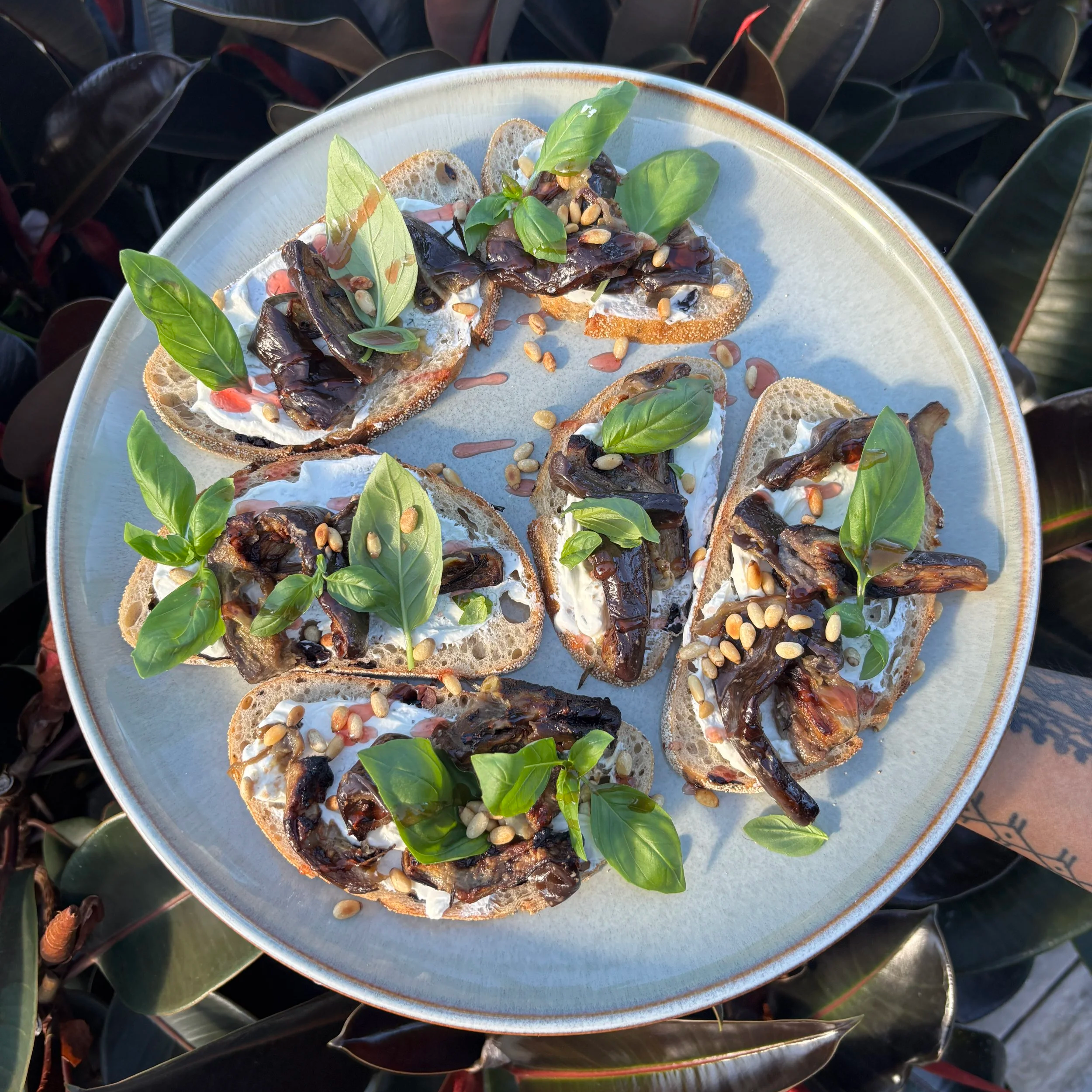 Close-up of a plate with bread slices topped with creamy cheese, roasted eggplant, fresh basil leaves, pine nuts, and drizzled with balsamic glaze.