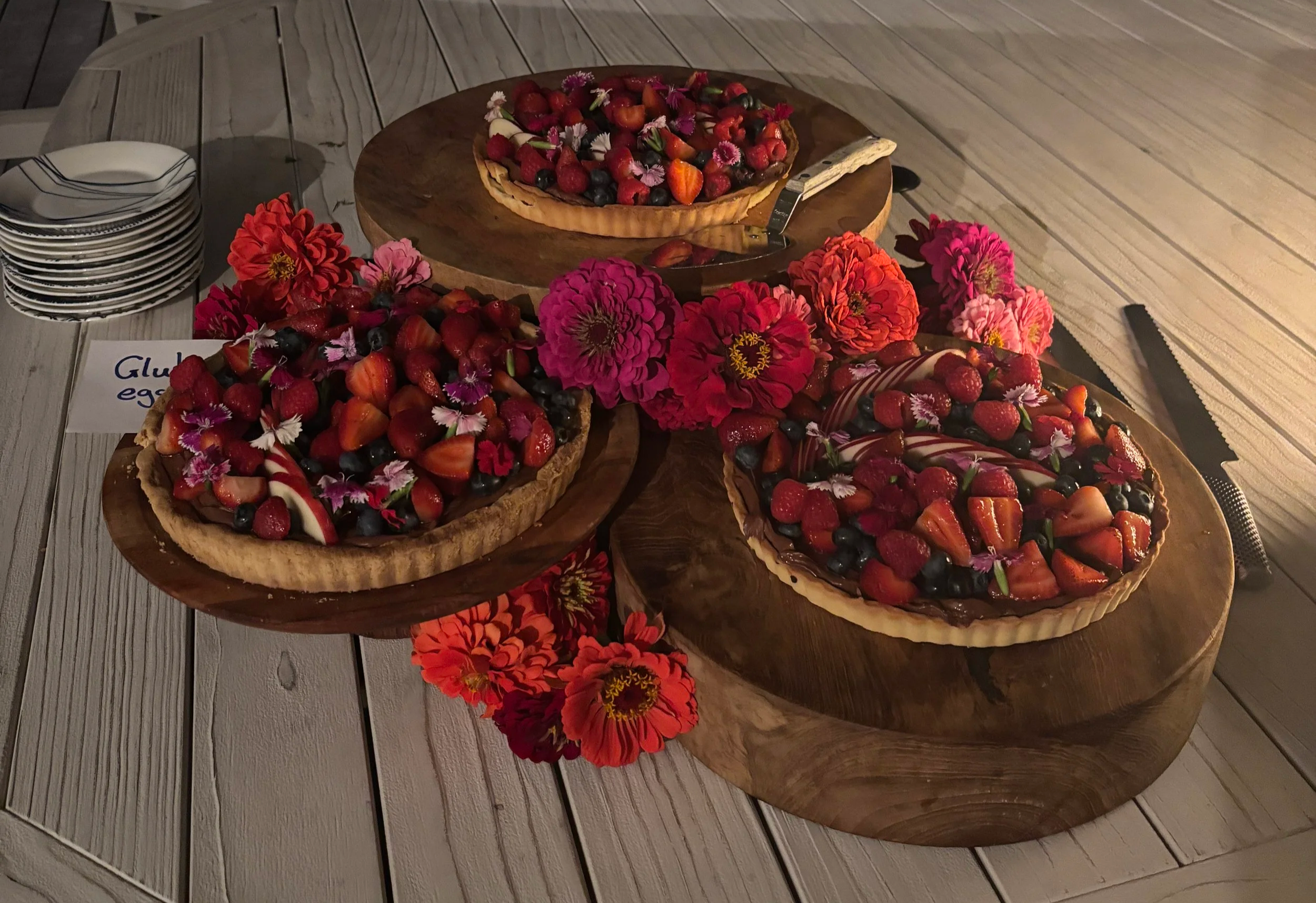Fruit tart topped with strawberries, blueberries, and edible flowers on a wooden board, surrounded by flowers and plates on a wooden table.