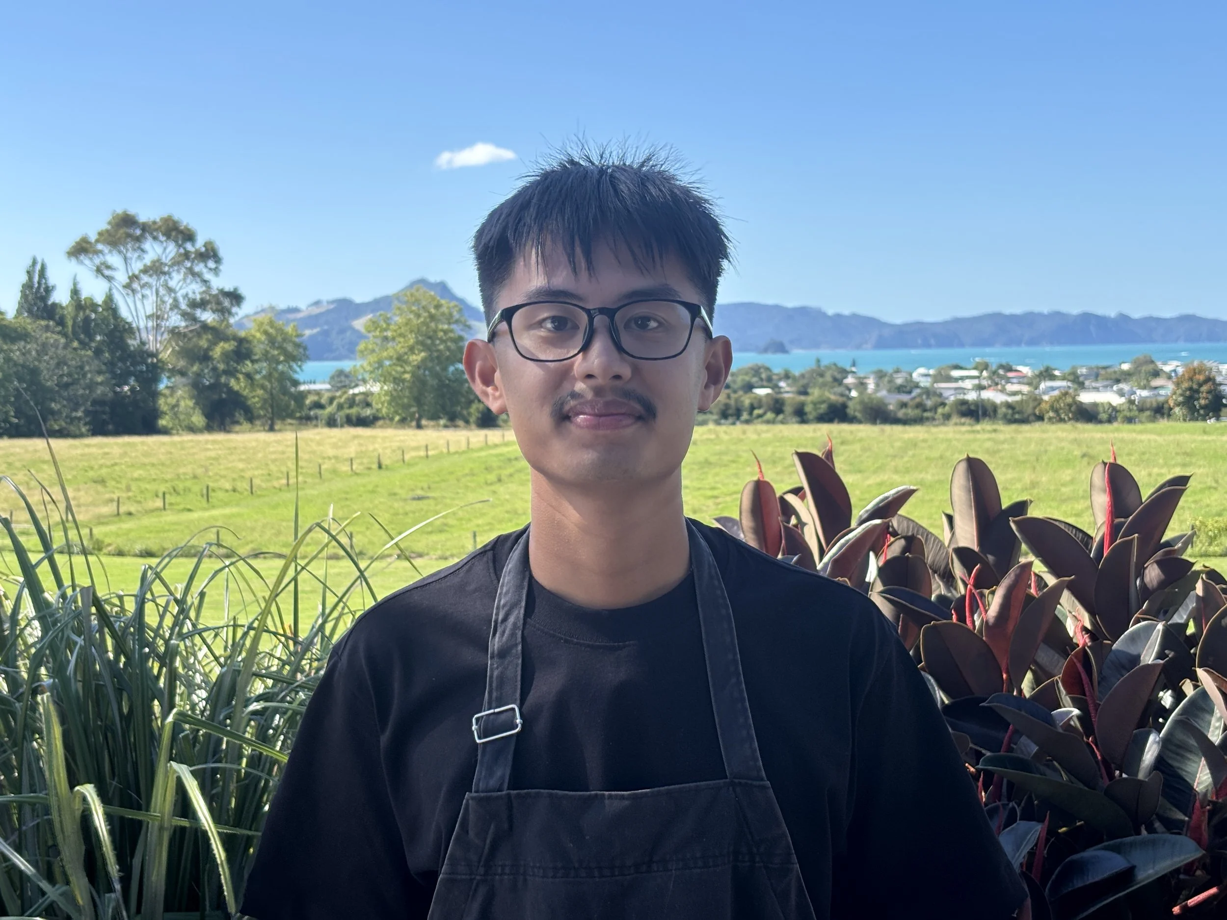 A young man with black hair, glasses, and a mustache standing outdoors with greenery, mountains in the background, and a lake or river visible behind him.