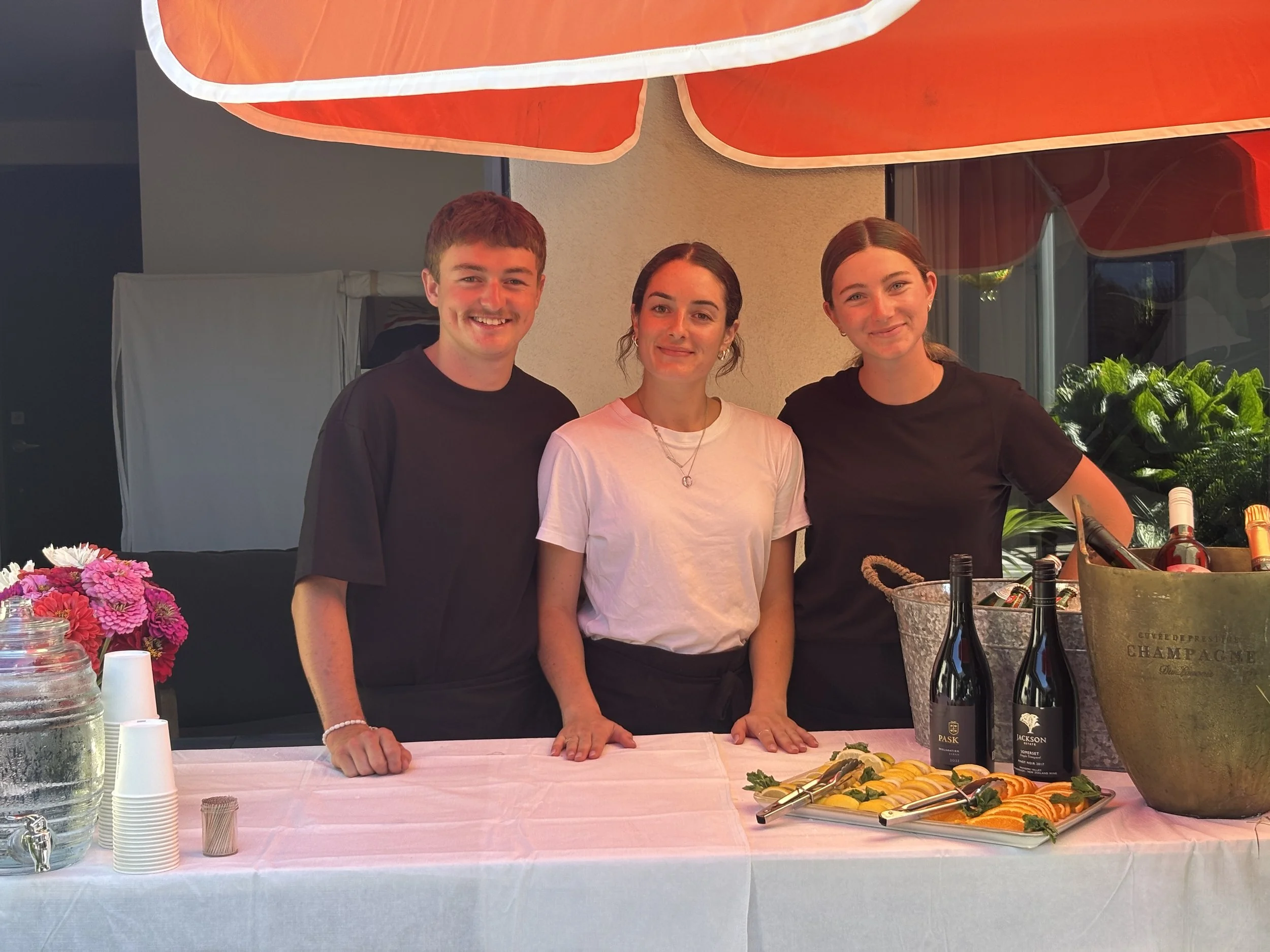 Three smiling young adults, two women and one man, stand behind a table with food and bottles of wine at an outdoor event under an orange umbrella.