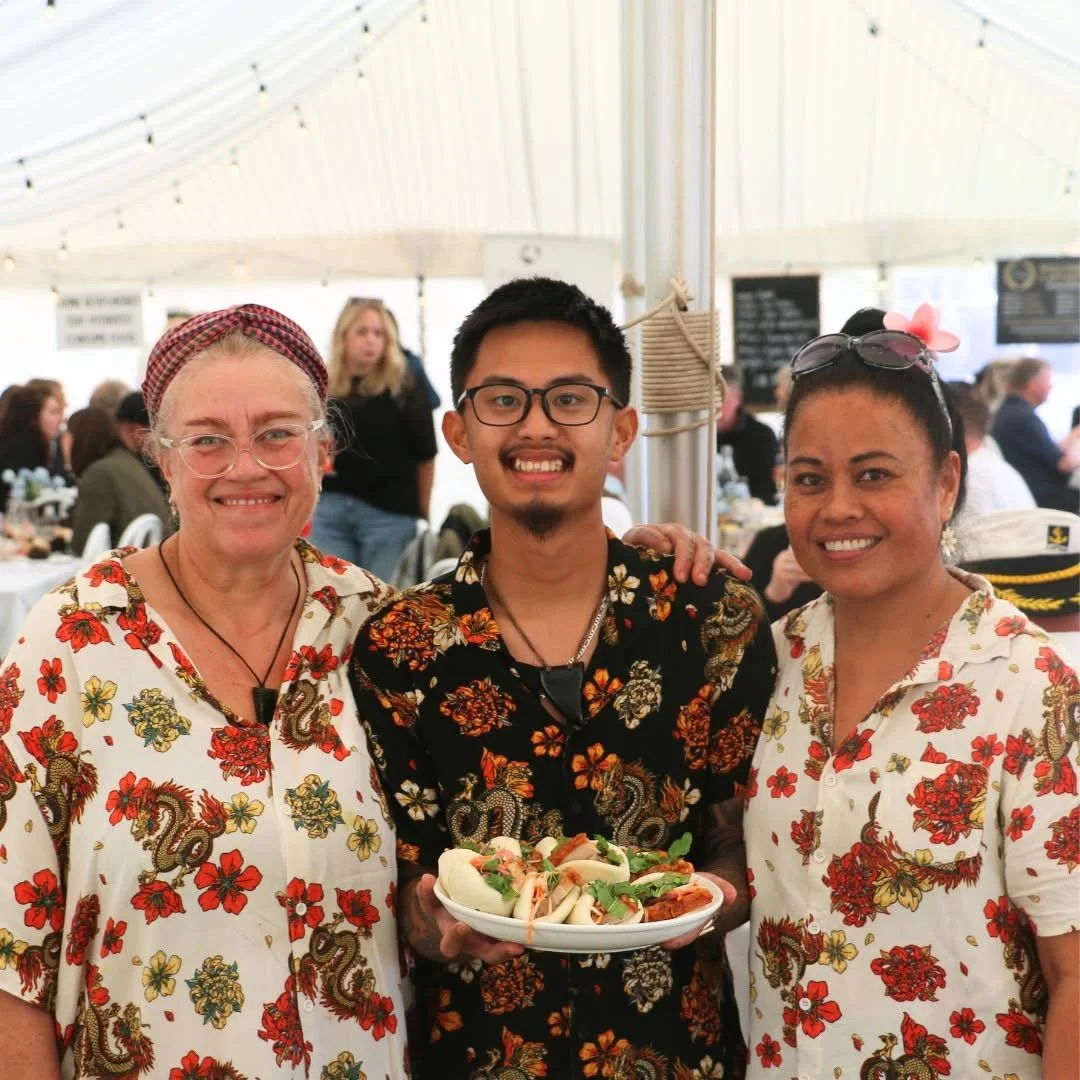 Three people smiling in a tent at a gathering, with two women on either side and a man in the middle holding a plate of food, all wearing floral shirts.