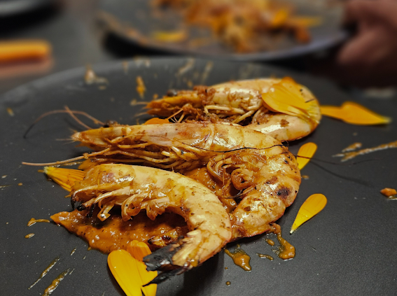 Three cooked shrimp on a black plate with yellow flower petals and sauce.