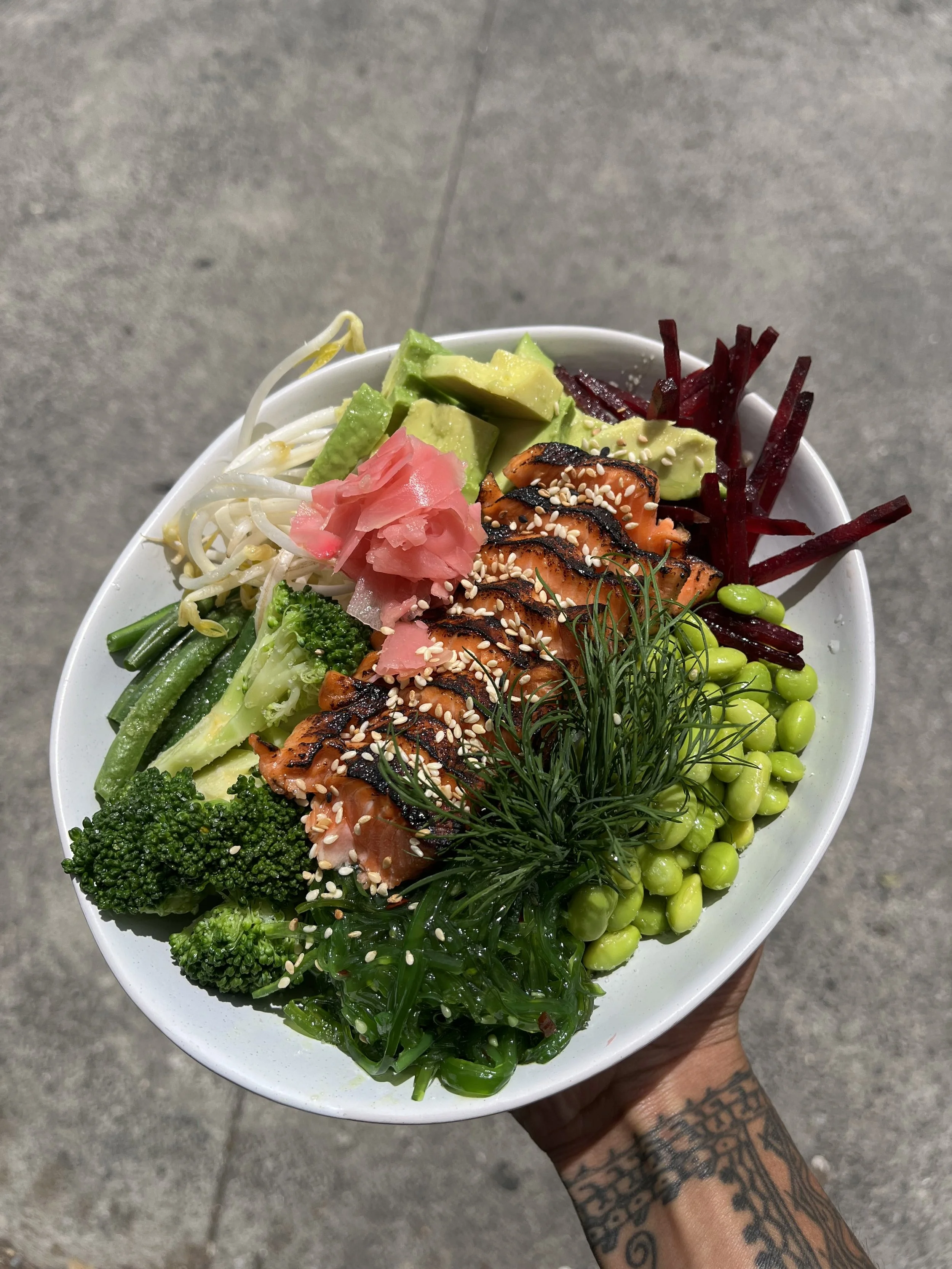 A bowl filled with assorted fresh vegetables including broccoli, edamame, bean sprouts, shredded beets, sliced avocado, grilled salmon with sesame seeds, and garnished with fresh herbs, all held by a tattooed hand on a concrete surface.