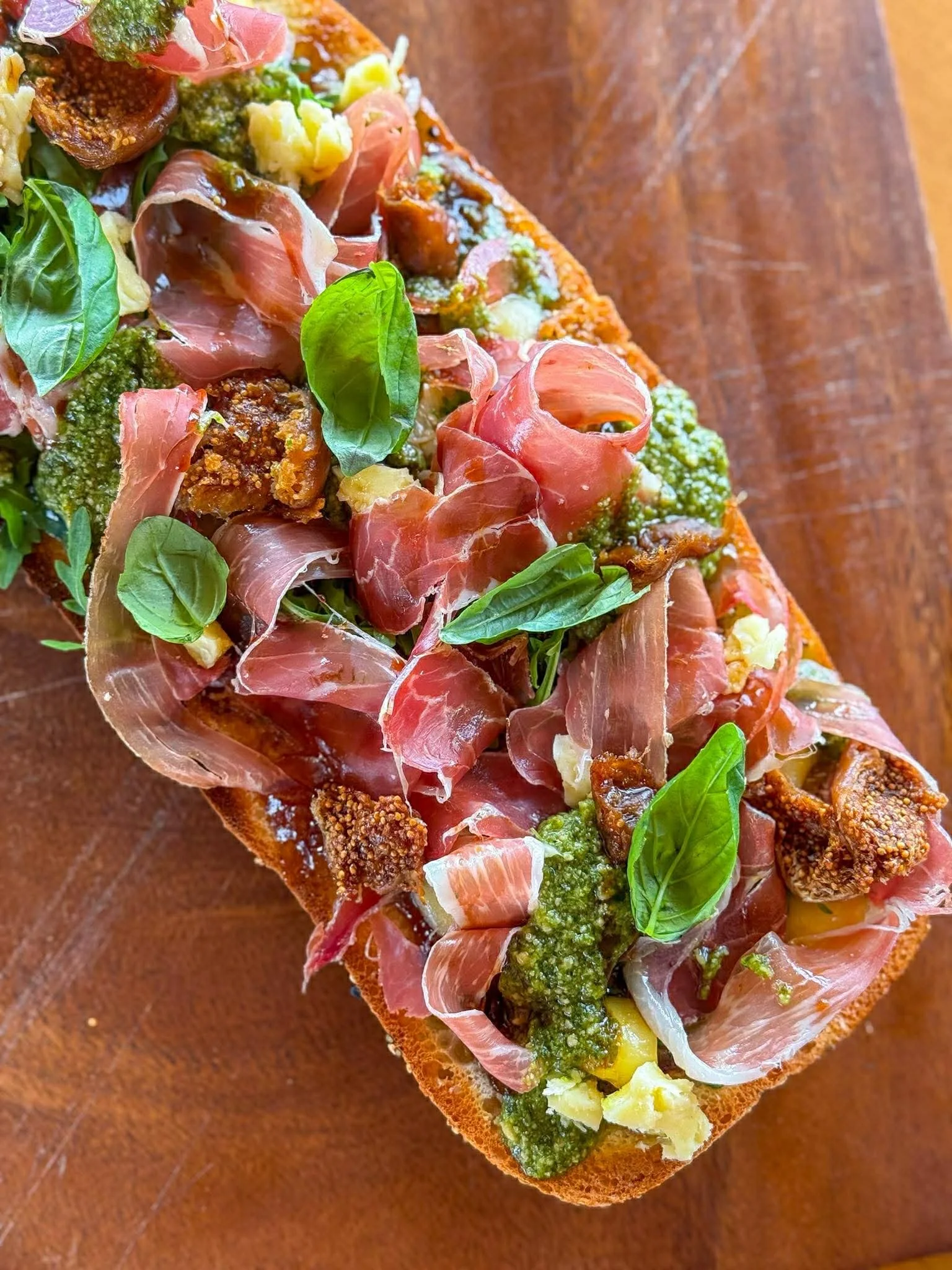 Close-up of a slice of bread topped with prosciutto, green pesto, fresh basil leaves, chopped hard-boiled egg, and sliced figs on a wooden surface.