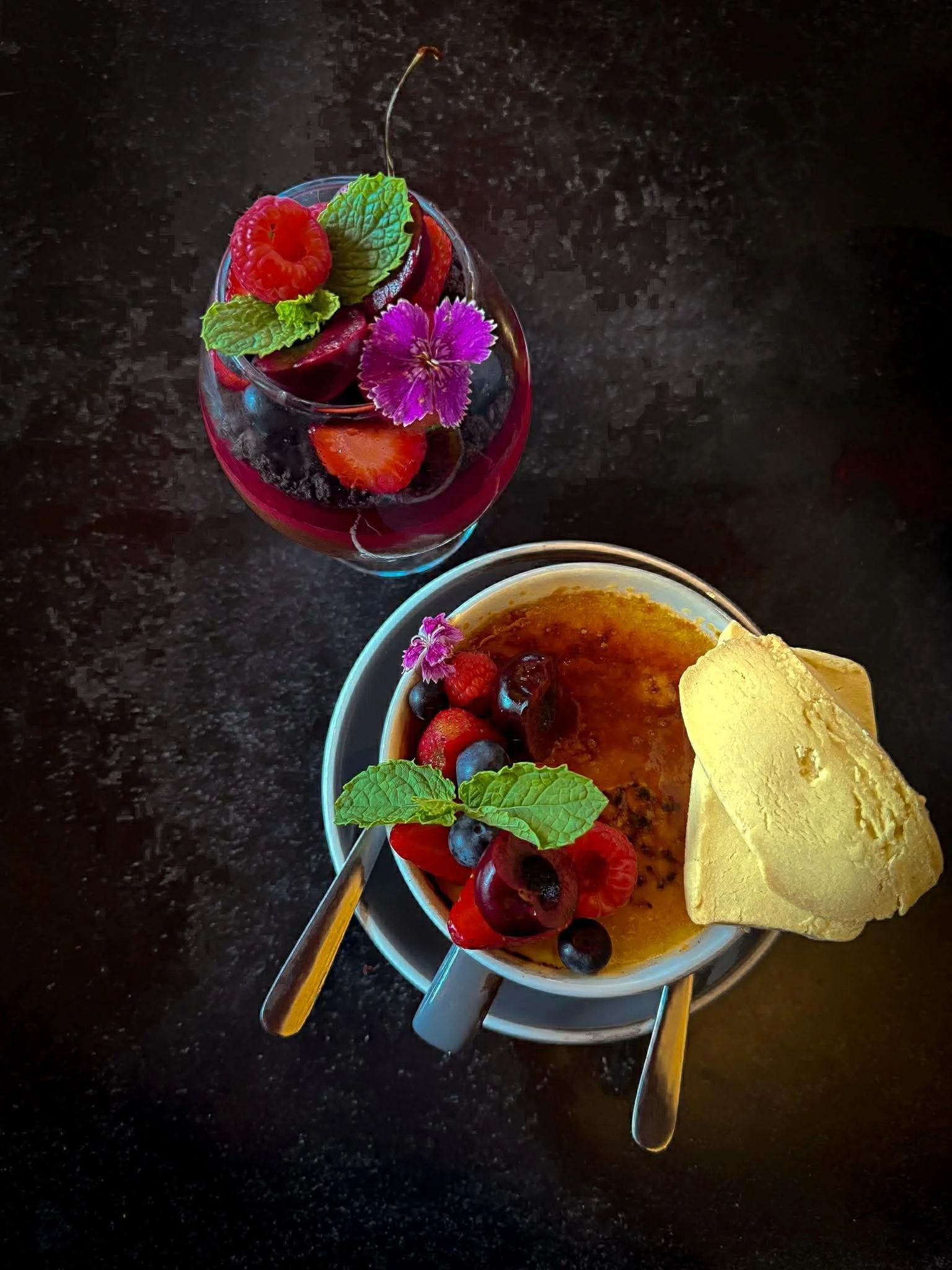 A glass of berry dessert garnished with fresh berries, mint leaves, and a purple flower, and a small bowl of crème brûlée topped with berries and edible flowers on a dark countertop.