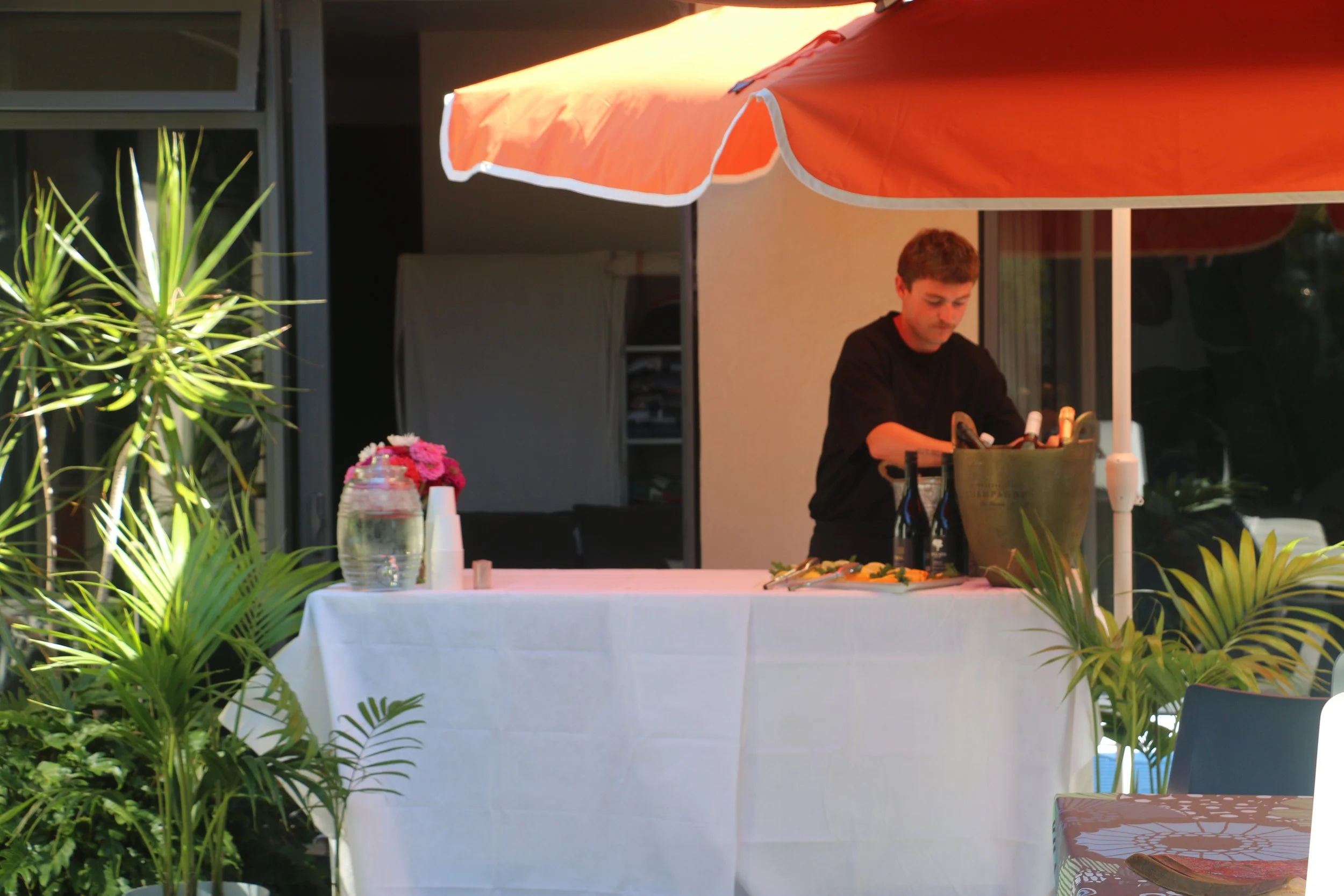 A man standing behind a table set for a meal outdoors, next to a large orange patio umbrella, with greenery in the foreground and flowers on the table.