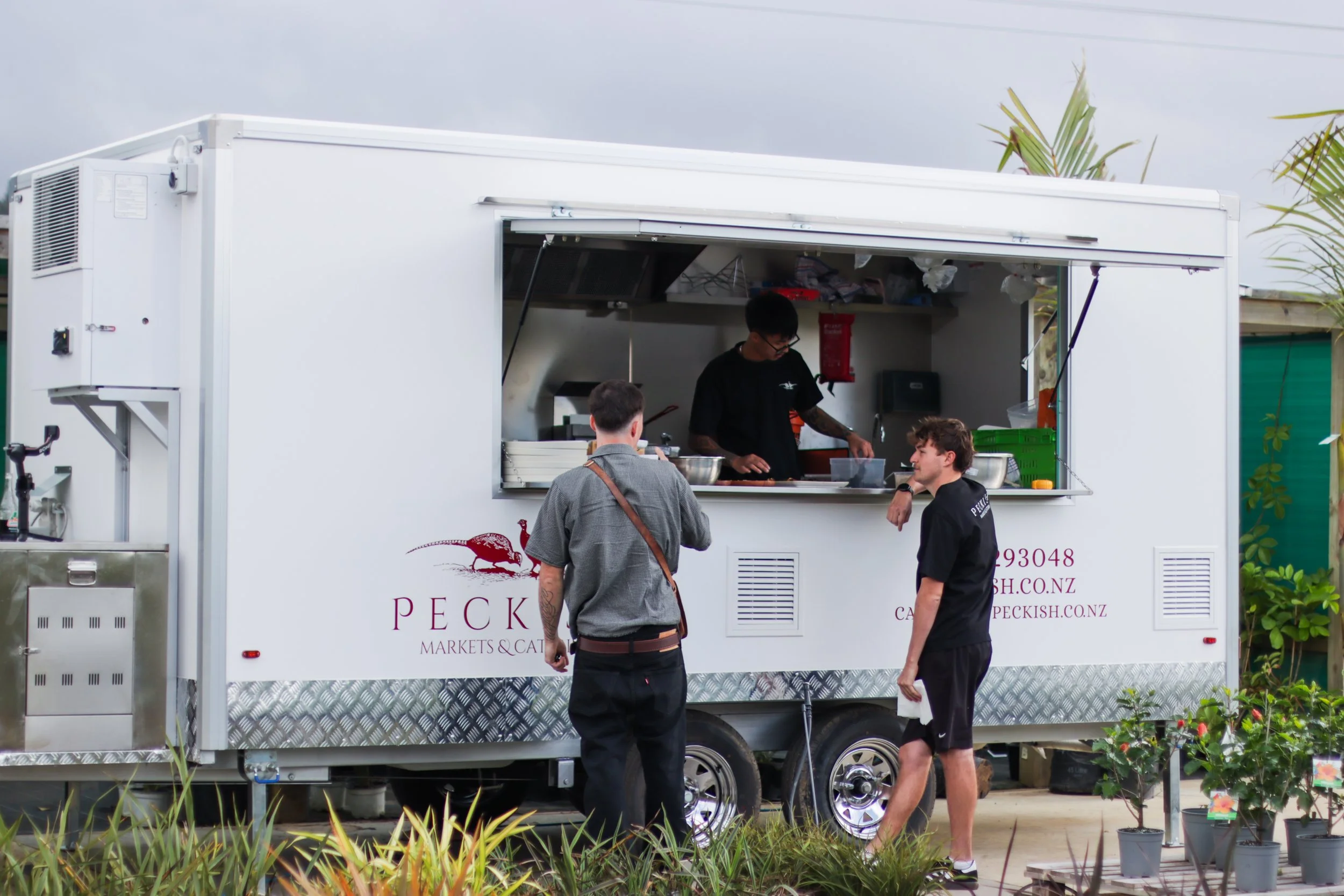 A white food truck with a large side window open, serving customers outdoors. Three people are interacting in front of the truck, with one inside preparing food. There are plants and a fence around the area.