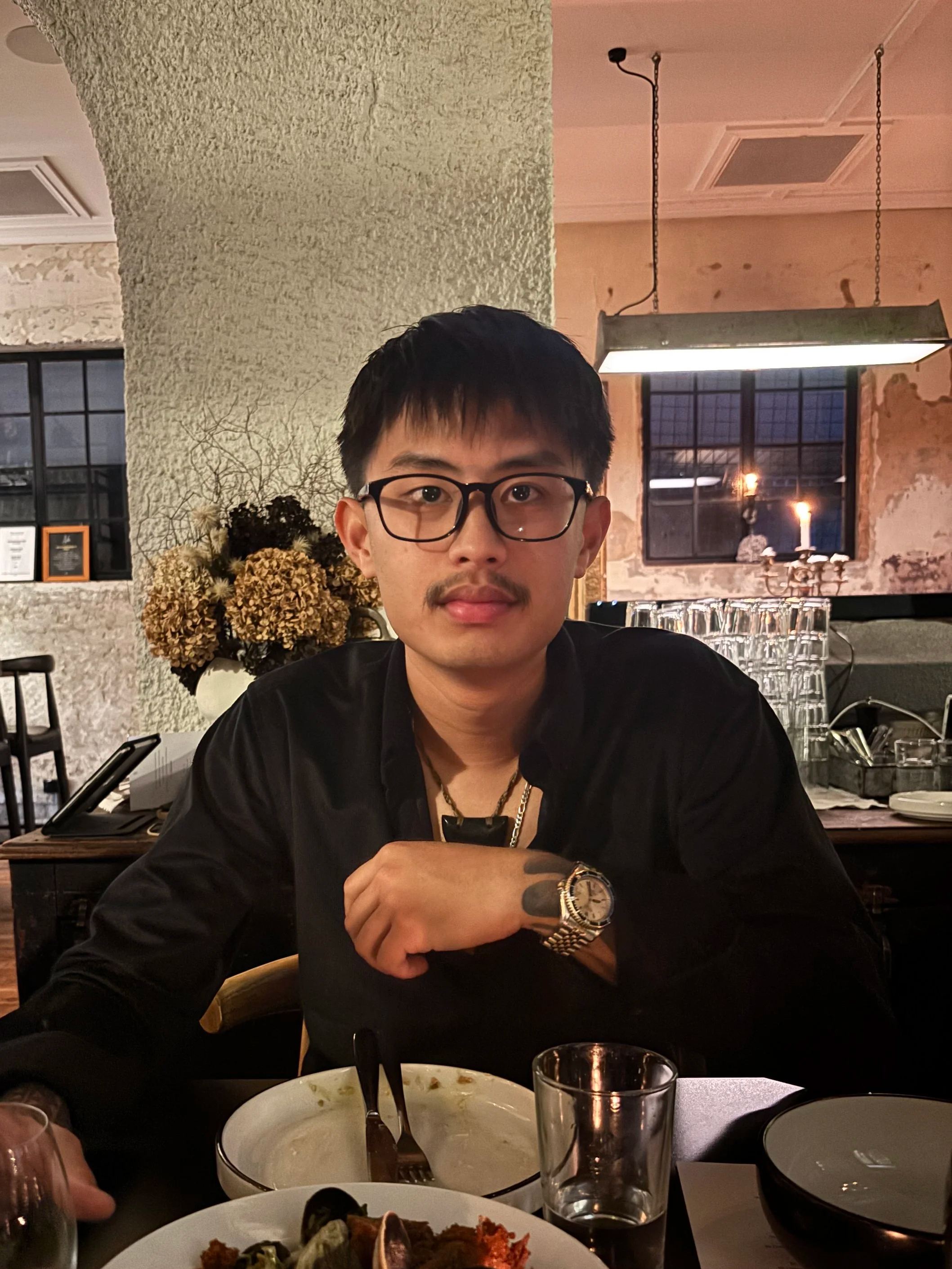 A man wearing glasses, a watch, and a necklace, sitting at a restaurant table with plates of food and glasses, with a rustic brick and wood interior, visible window, and decorative candles in the background.