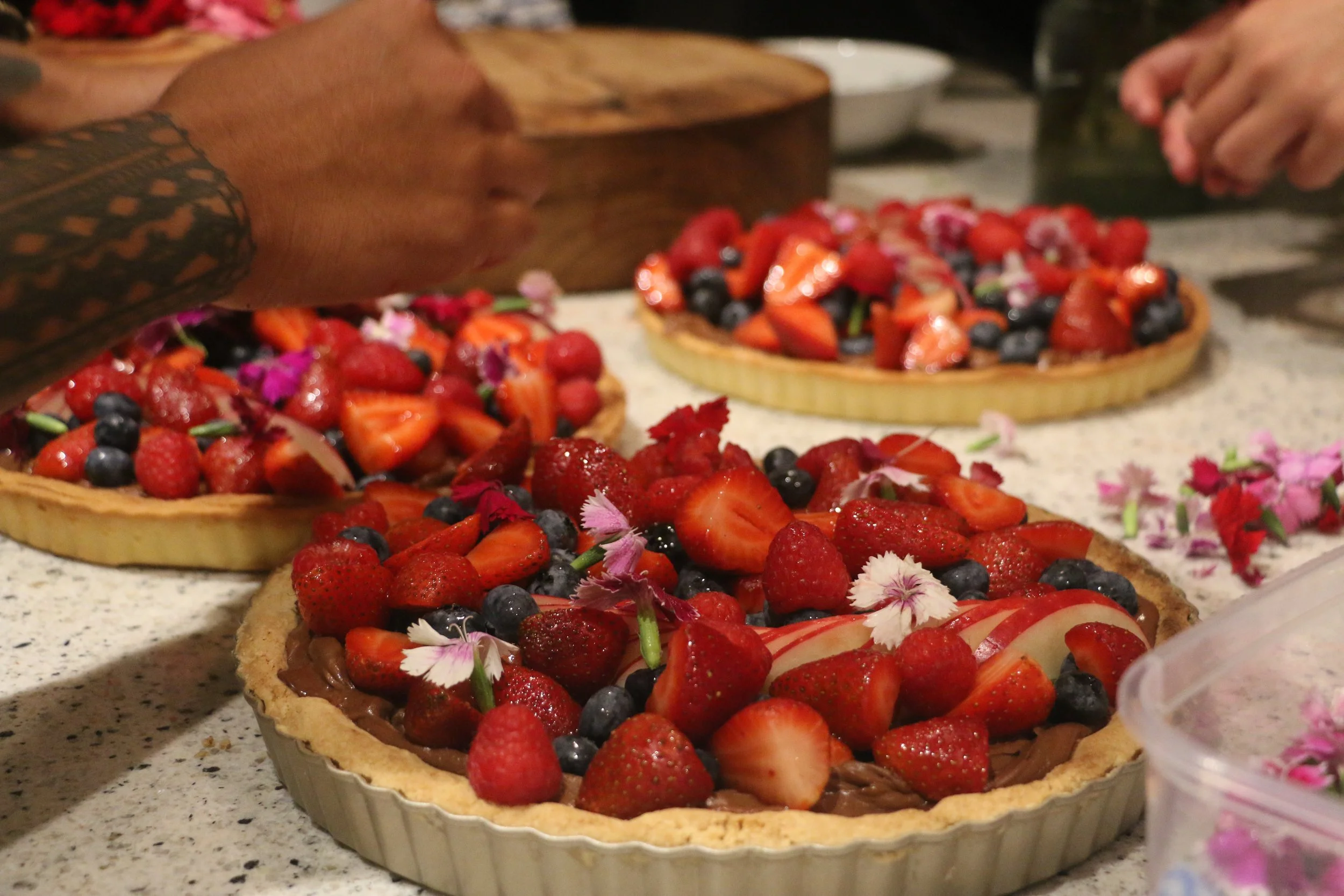 Close-up of a fruit tart topped with strawberries, blueberries, and edible flowers on a speckled countertop.
