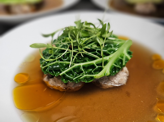 Gourmet dish with a piece of meat topped with a leafy green vegetable and microgreens, served in a brown sauce