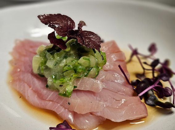 Sliced raw fish topped with chopped green onions and garnished with purple-leaf microgreens, served with a side of yellow sauce on a white plate.