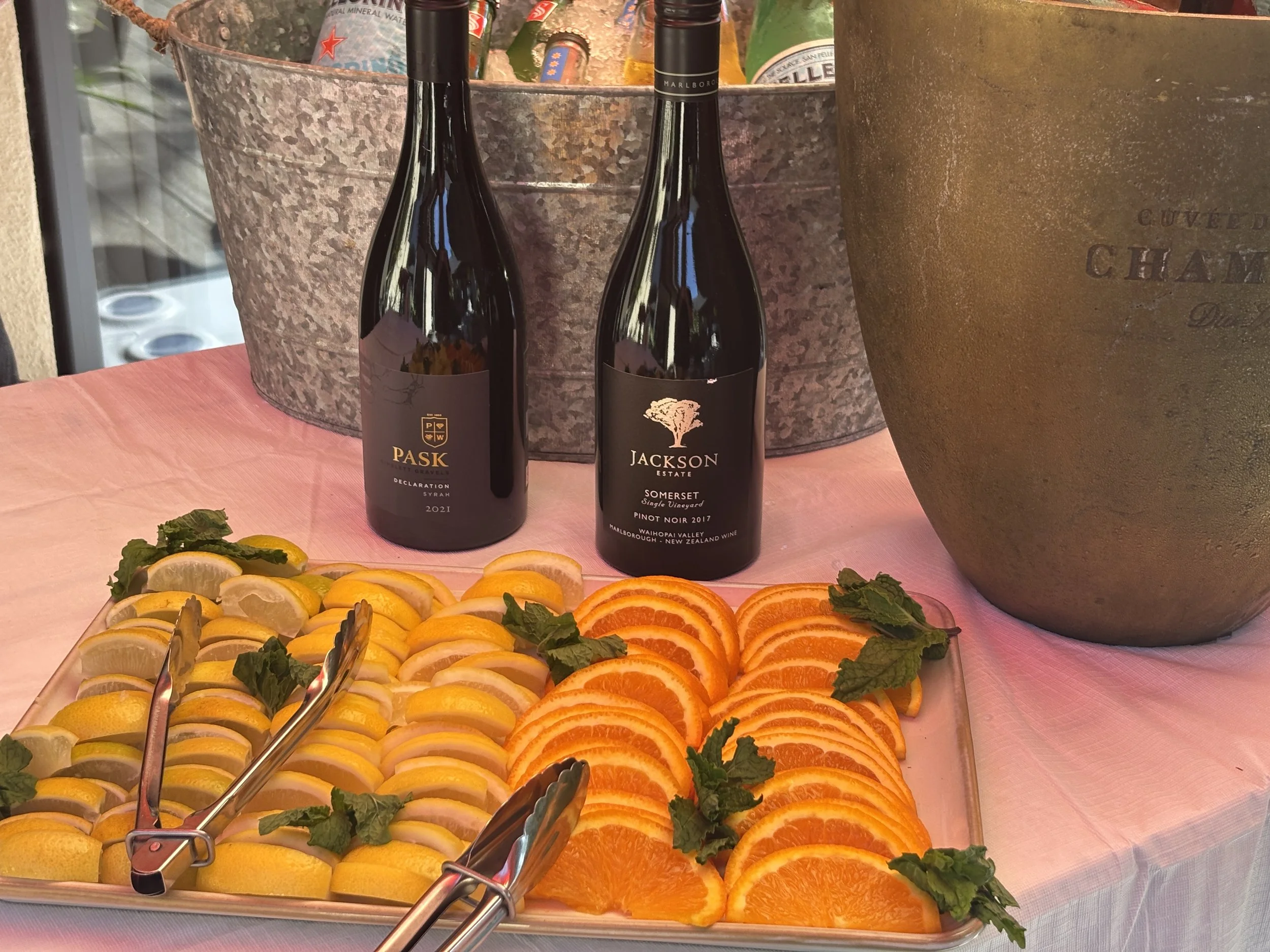 A display of sliced citrus fruits including lemons, oranges, and grapefruits arranged on a tray with some mint leaves. Behind the fruits, there are two bottles of wine, one labeled 'Pask' and the other 'Jackson Estate,' with a large, rust-colored vas