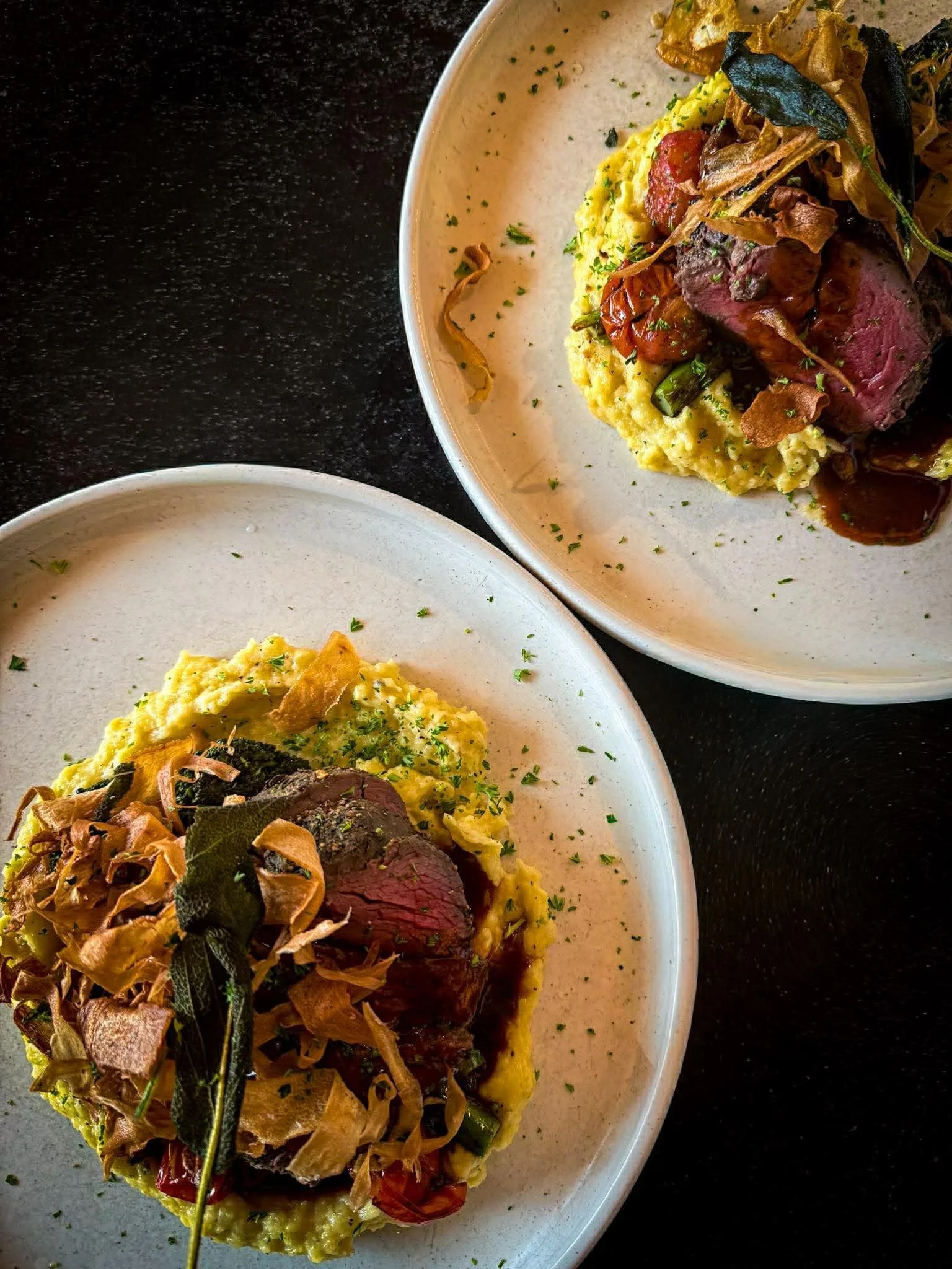 A plate of food featuring a meat dish with herbs on top, served with a bed of creamy yellow sauce, garnished with crispy fried onions and fresh herbs. Another plate with scrambled eggs garnished with herbs is partially visible in the foreground.