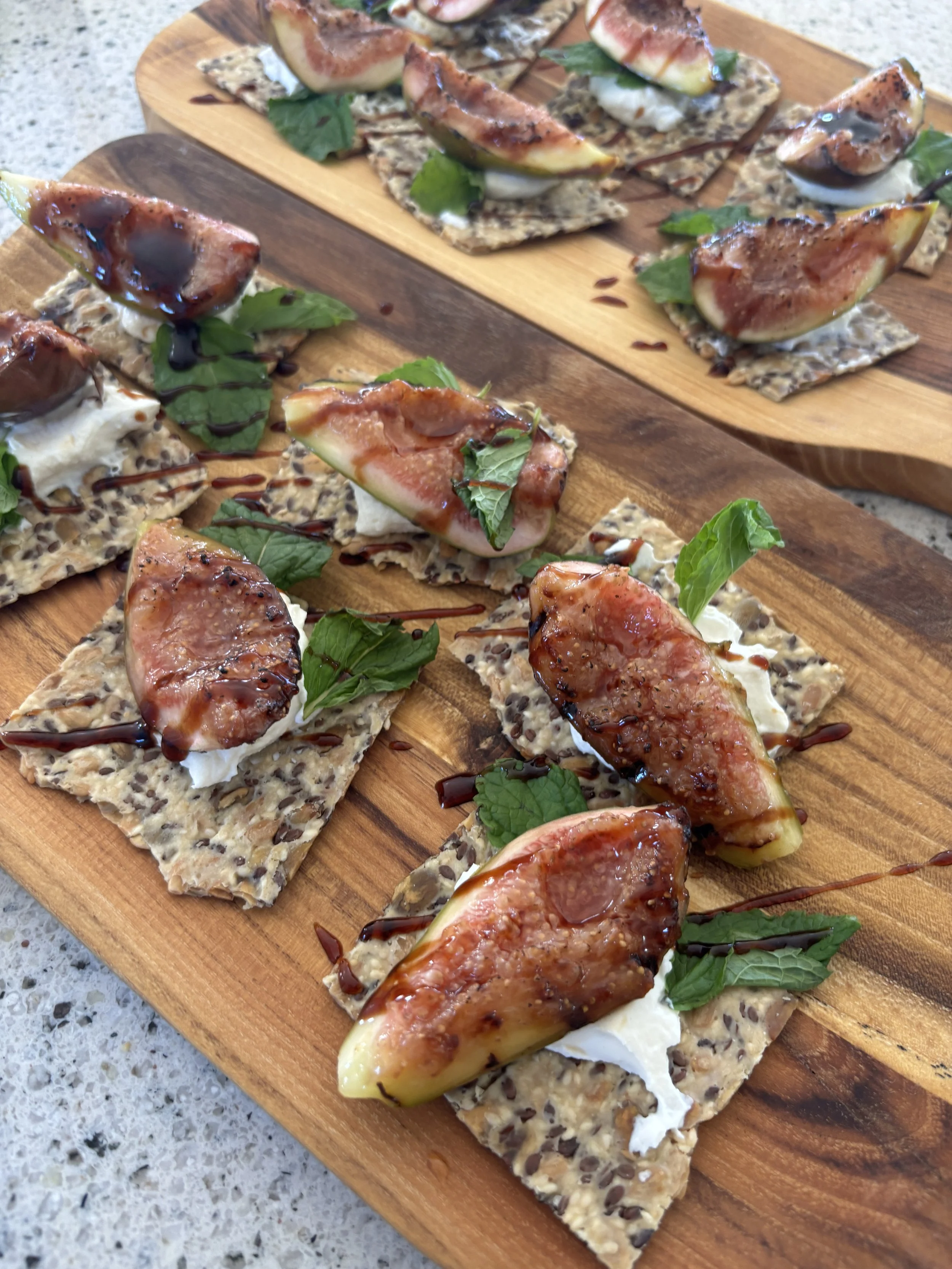 Wooden platter with crispbreads topped with ricotta cheese, fresh figs, fresh mint leaves, and balsamic glaze.