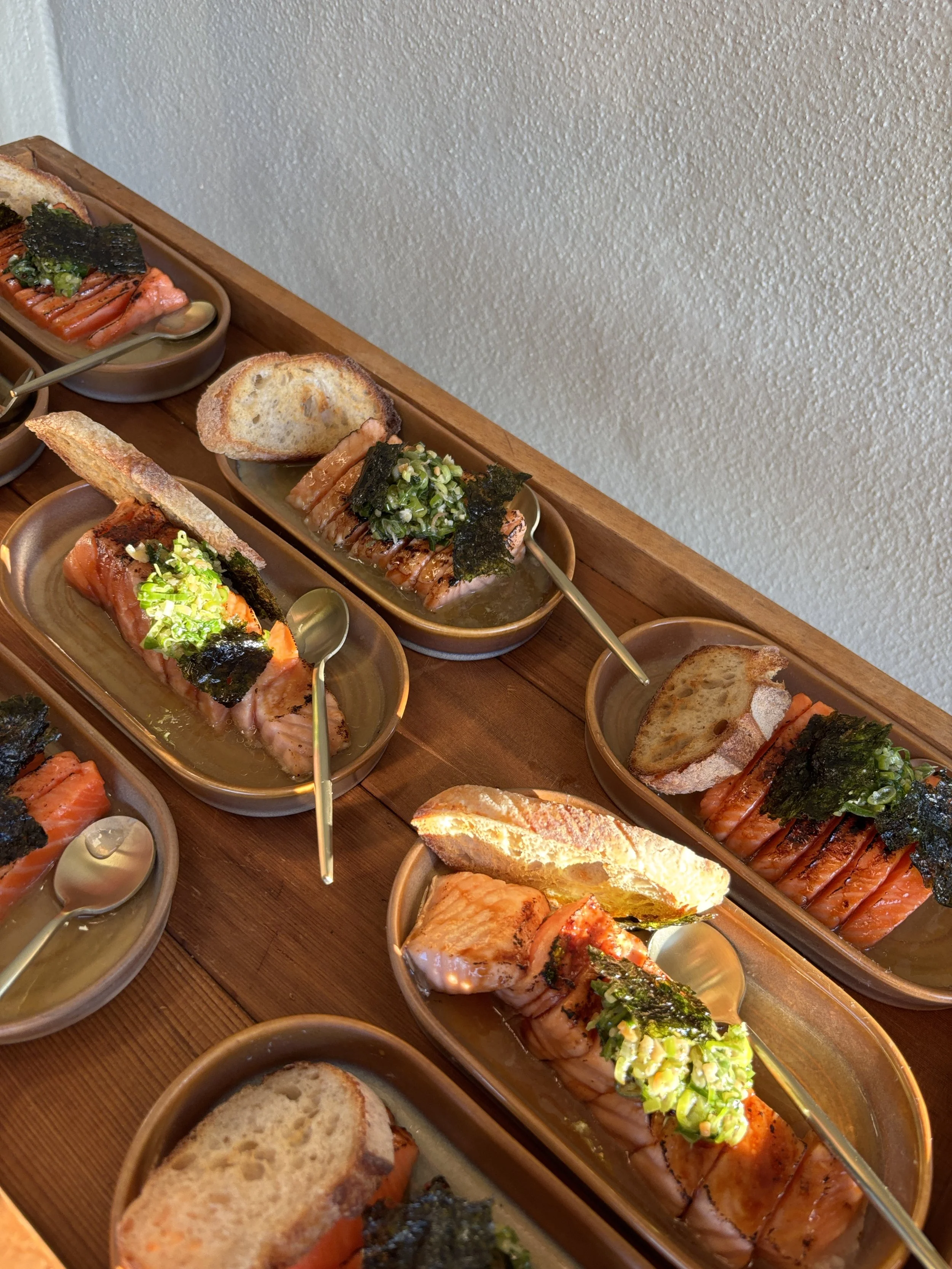 Close-up of multiple glazed salmon fillets topped with chopped green onions and seaweed, served on small brown plates with slices of bread, on a wooden table.