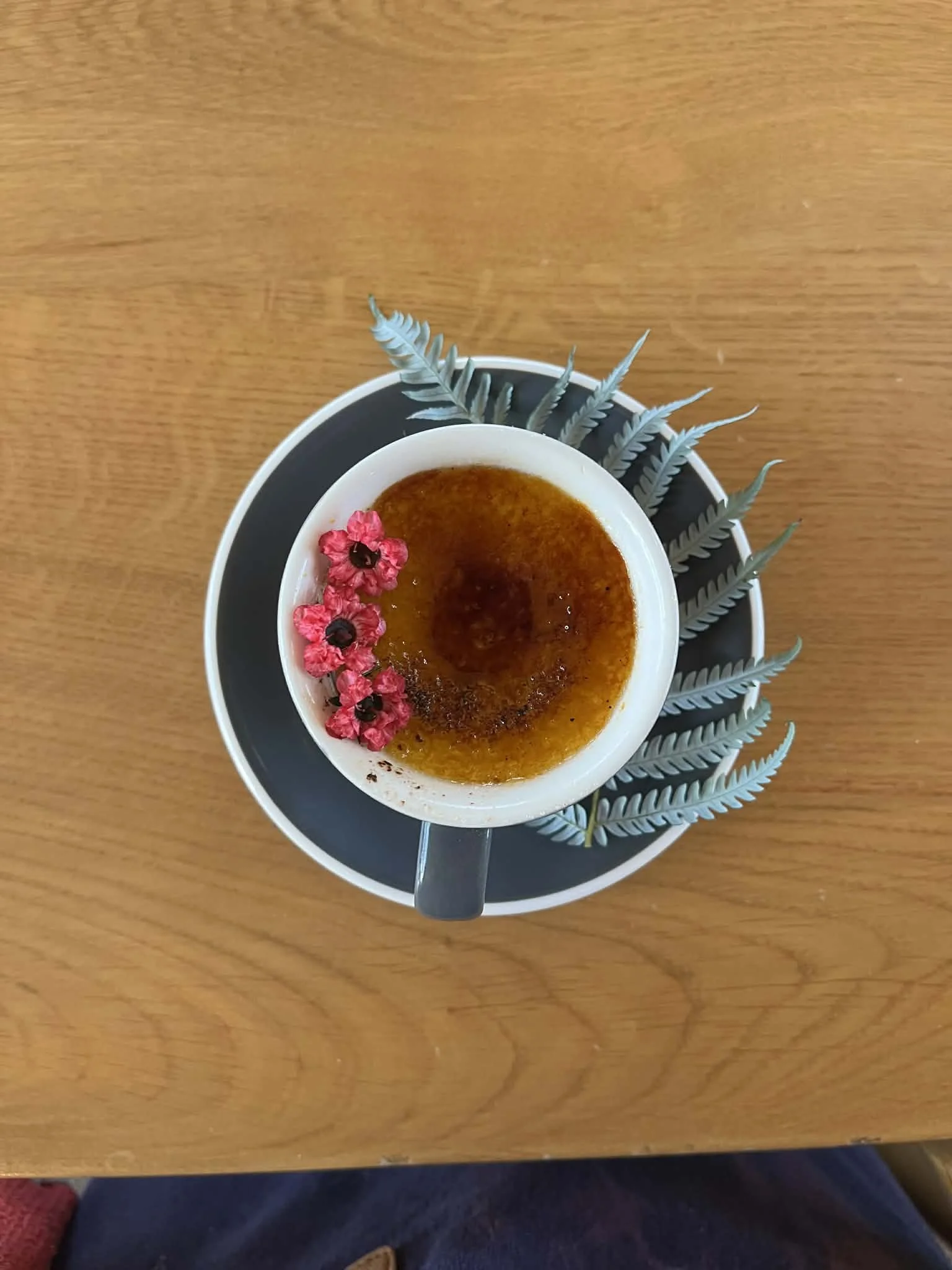 A cup of caramelized desserts garnished with pink flowers and black berries, placed on a black plate with gray leaves, on a wooden table.