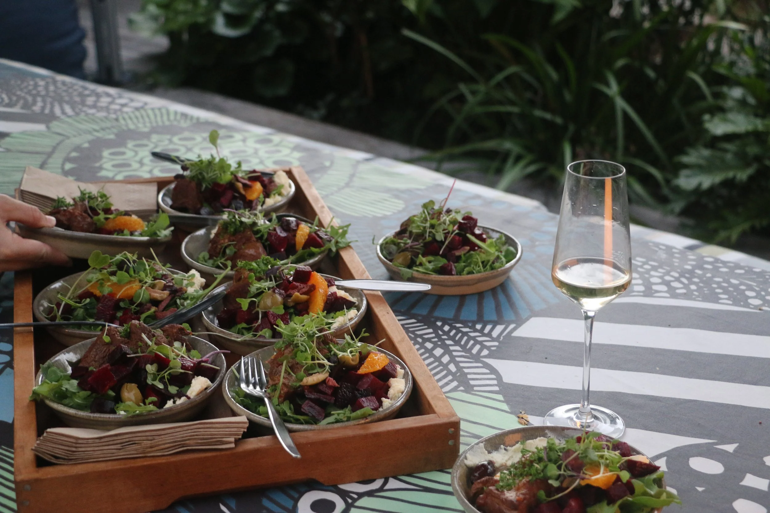 Table with multiple bowls of colorful salads and a glass of white wine, outdoors with greenery in the background.