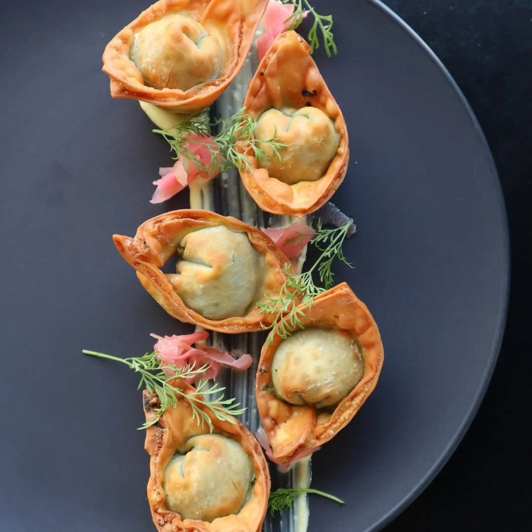 Five baked stuffed pepper halves with melted cheese, garnished with fresh herbs and pink pickled ginger on a black plate.