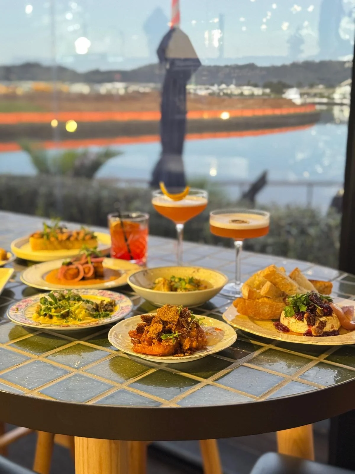 Table with various plates of food and three cocktails near a window overlooking a waterfront scene with industrial structures and distant hills.