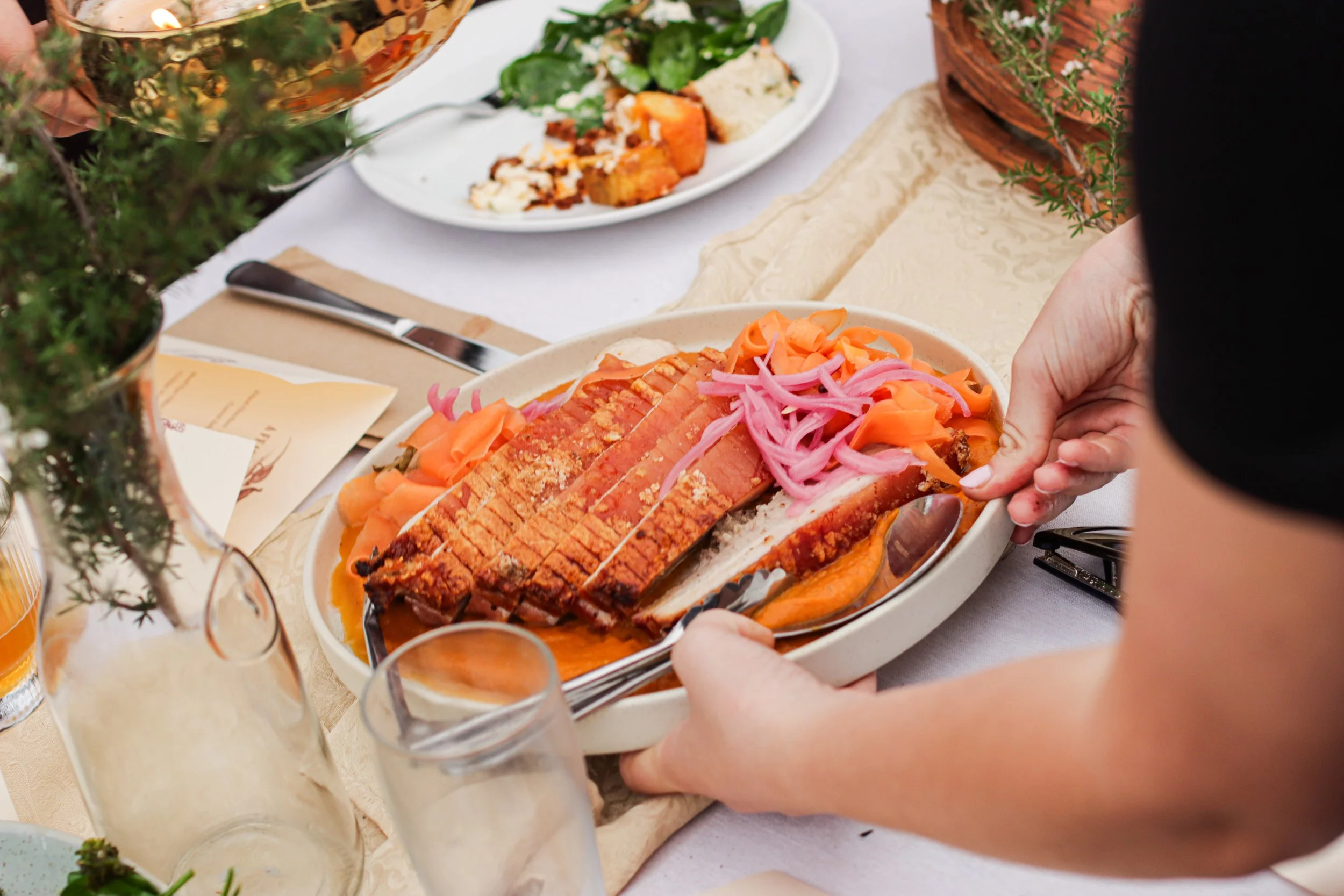 Person serving sliced roasted meat with pickled onions on a white platter at a dining table.