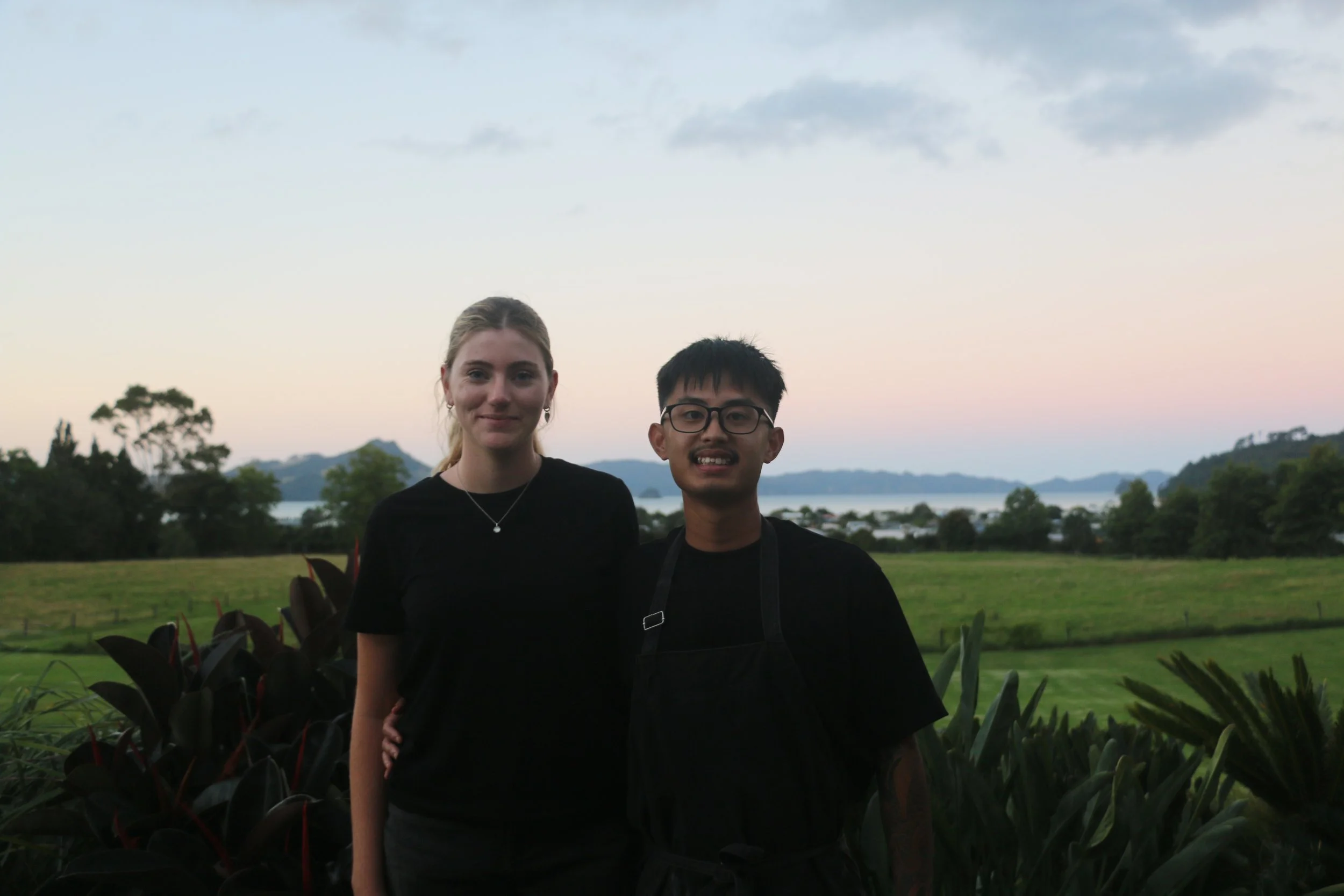 A young woman and a young man standing outdoors during sunset with a scenic landscape of trees, water, and mountains in the background.