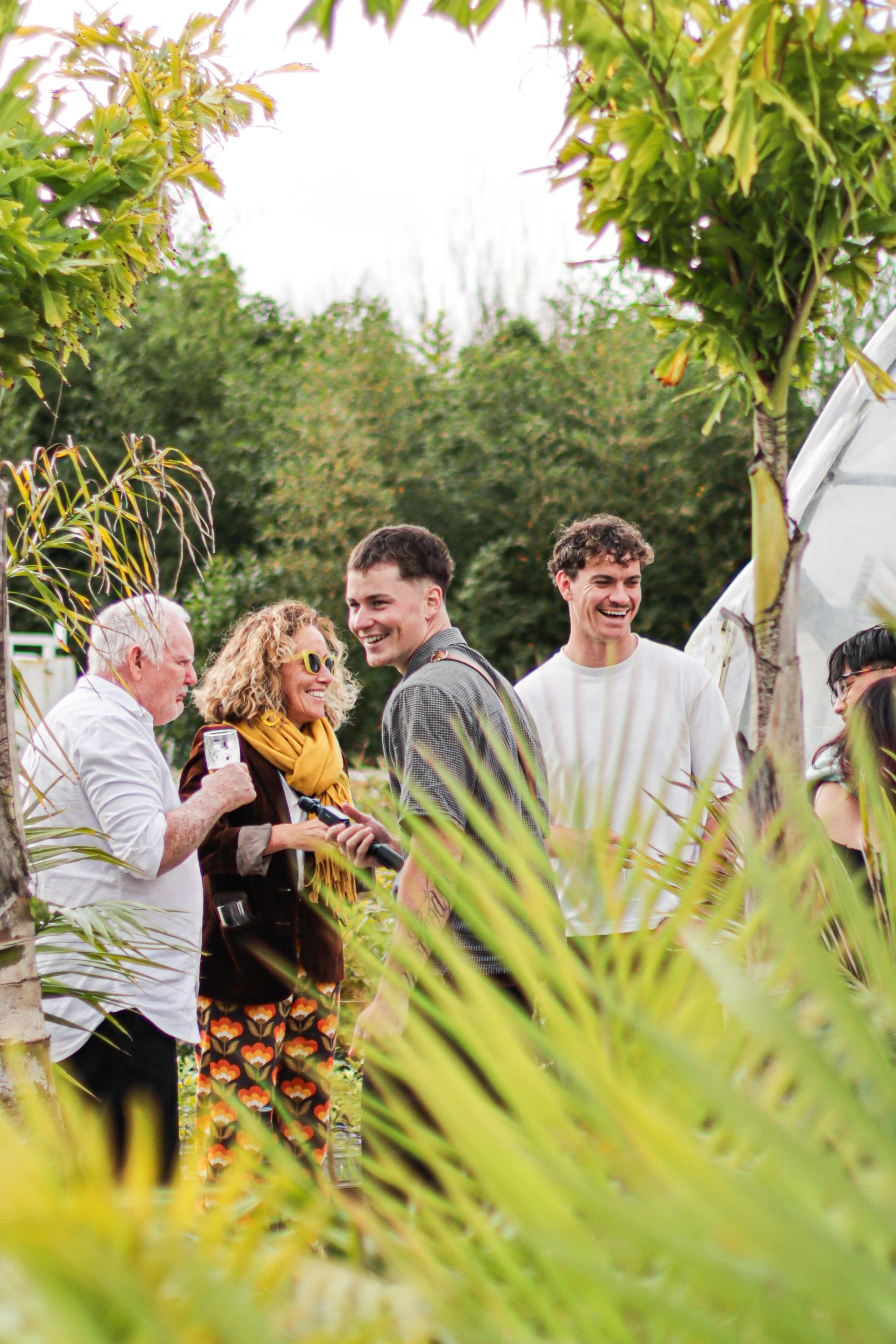 A group of people standing outdoors, smiling and talking among trees and plants.