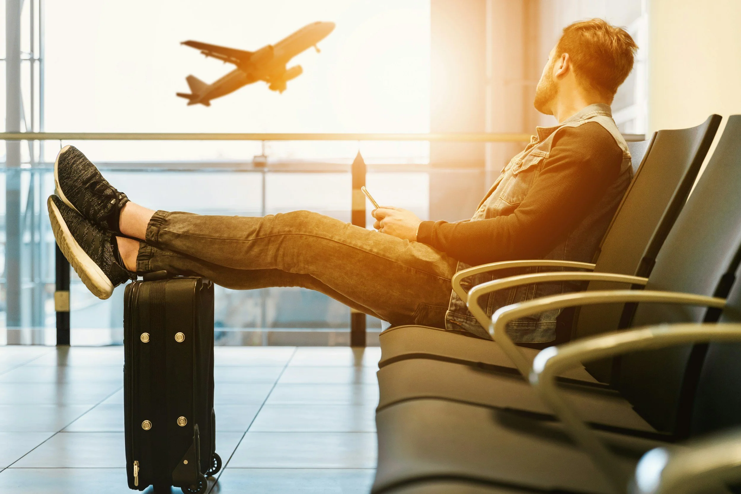 A man sitting in an airport waiting area, looking at an airplane flying outside the large window.