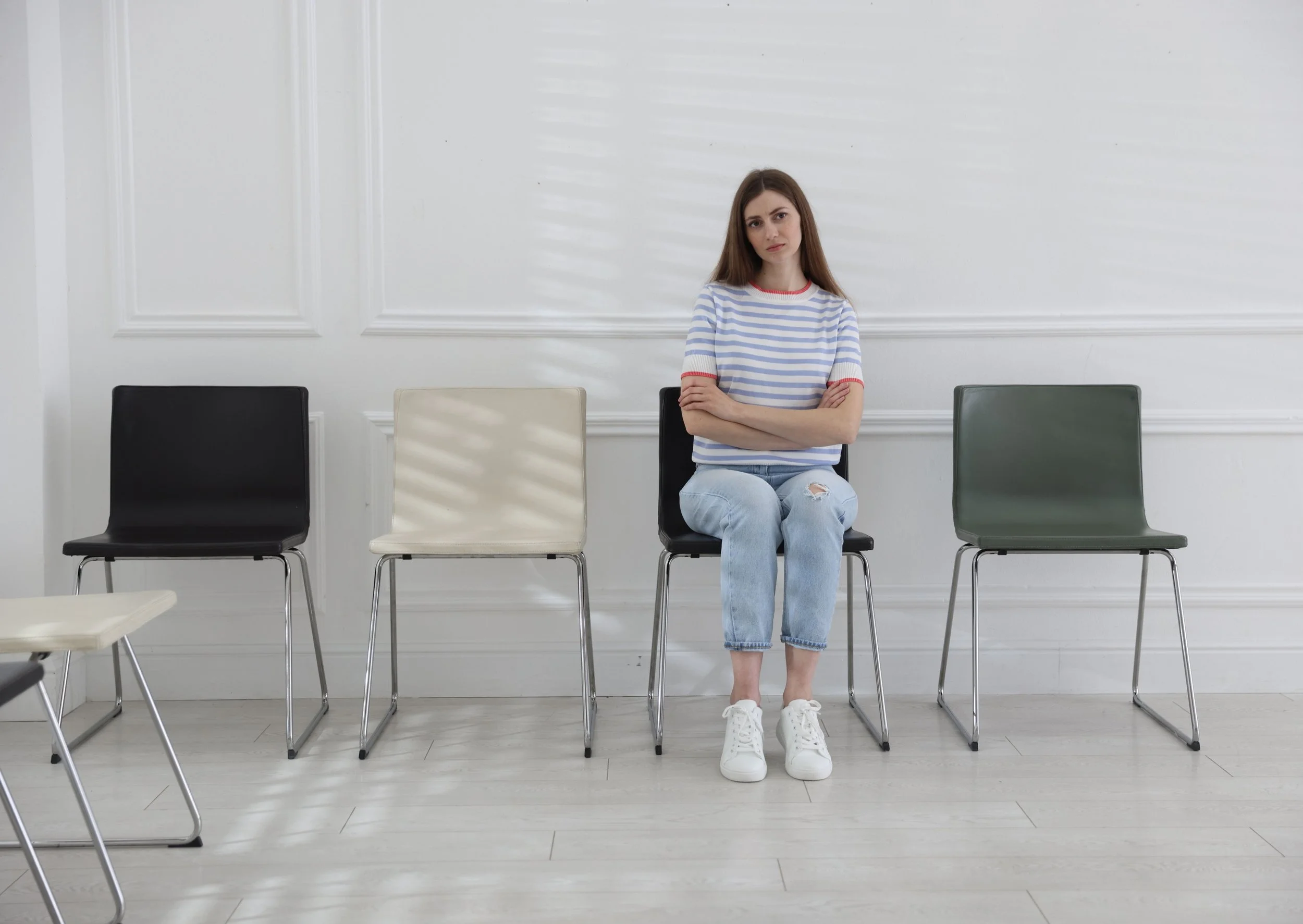 A young woman with long brown hair, wearing a striped t-shirt and ripped jeans, sitting alone on a black chair in a healthcare waiting room with white walls and light-coloured wooden flooring, surrounded by empty chairs.