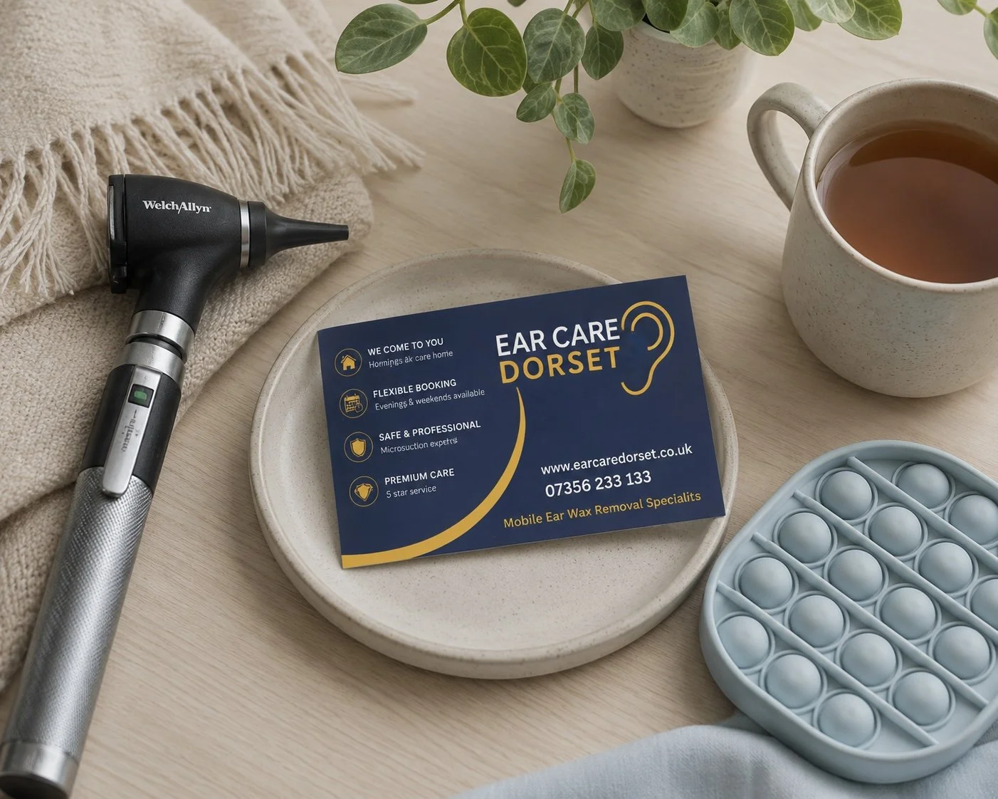 A business card for Ear Care Dorset, with a digital otoscope, a cup of tea, a teal silicone bubble pop fidget toy, and a plant on a light wood surface.