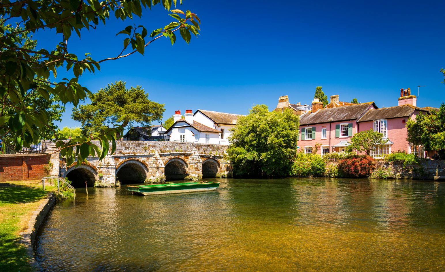 A picturesque scene of Christchurch Quay. A stone bridge with arches over a tranquil river, lined with charming houses and lush green trees under a bright blue sky.