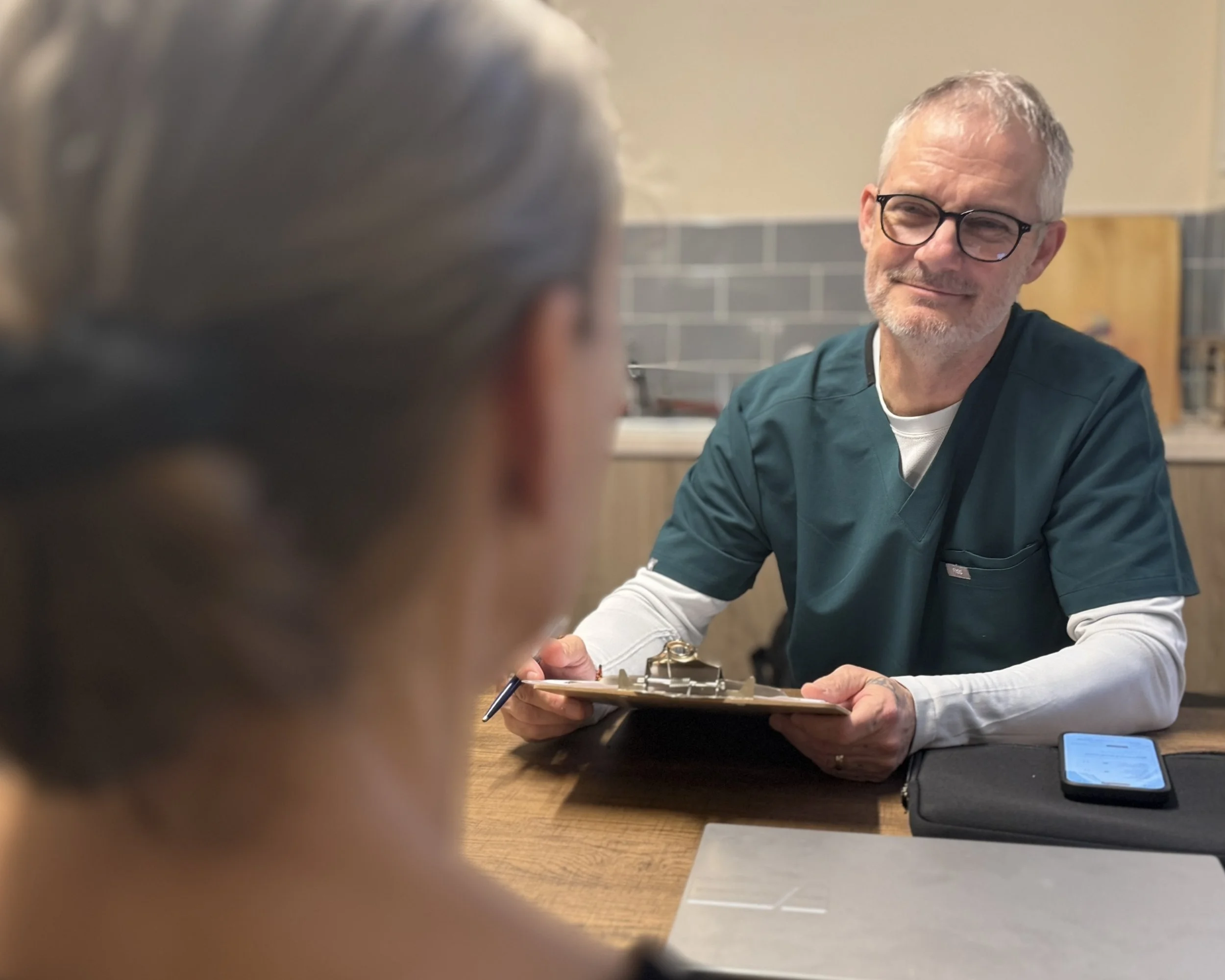 Our practitioner, Alan, smiling while sitting at a table, holding a clipboard with papers, completing an pre-treatment assessment with a person in the foreground out of focus.