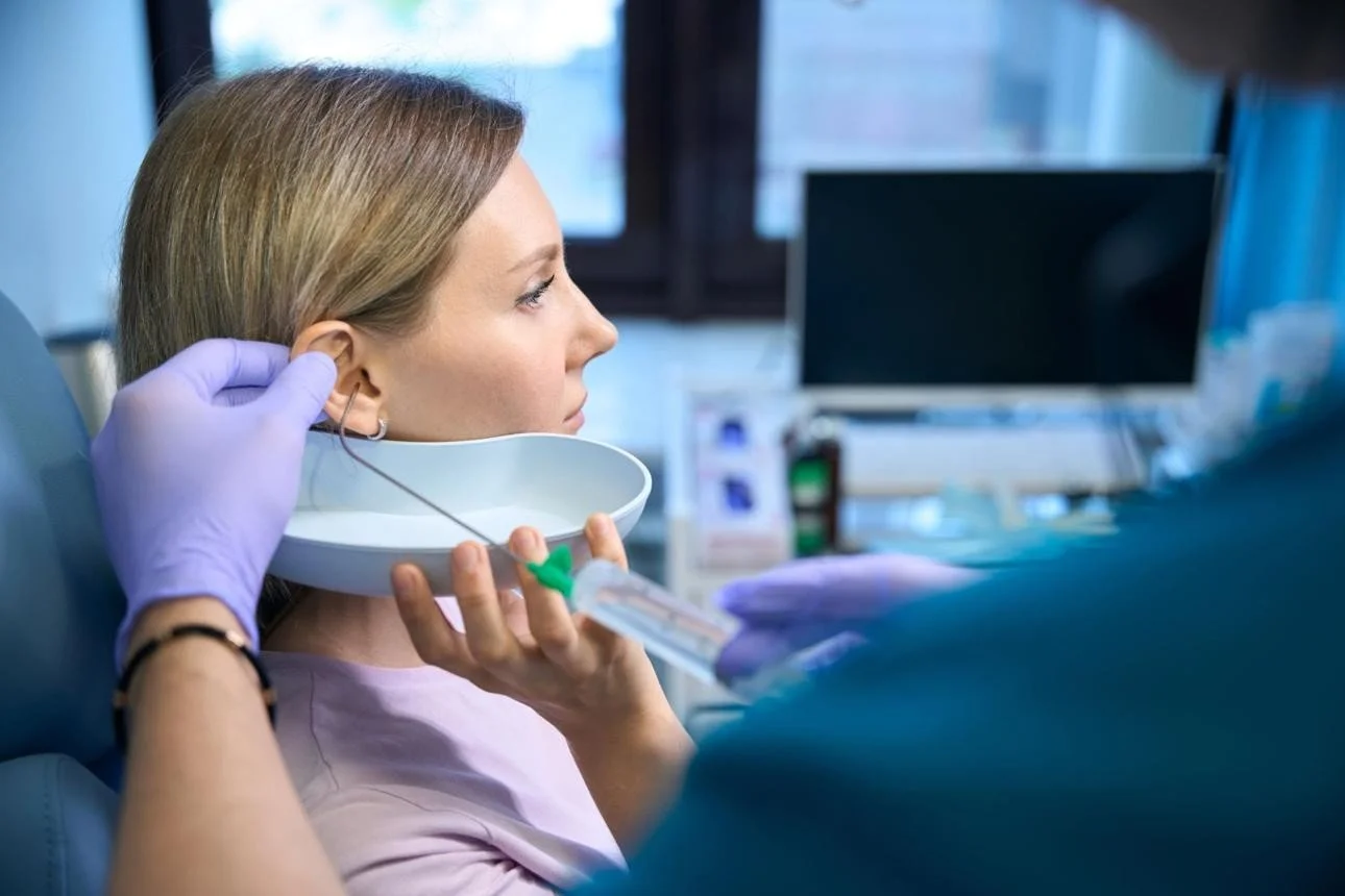 A woman with light brown hair receives ear irrigation treatment treatment from a professional ear wax removal practitioner wearing medical gloves.