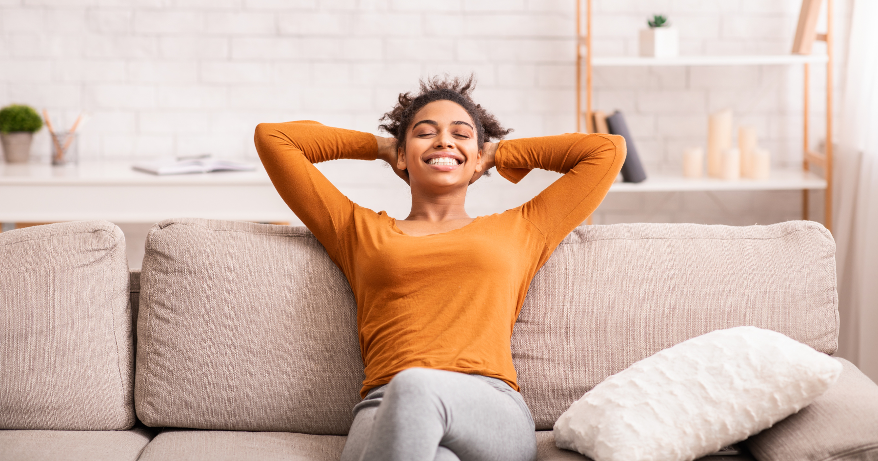 A smiling woman relaxing on a sofa with her hands behind her head, sitting in a comfortable living room setting.